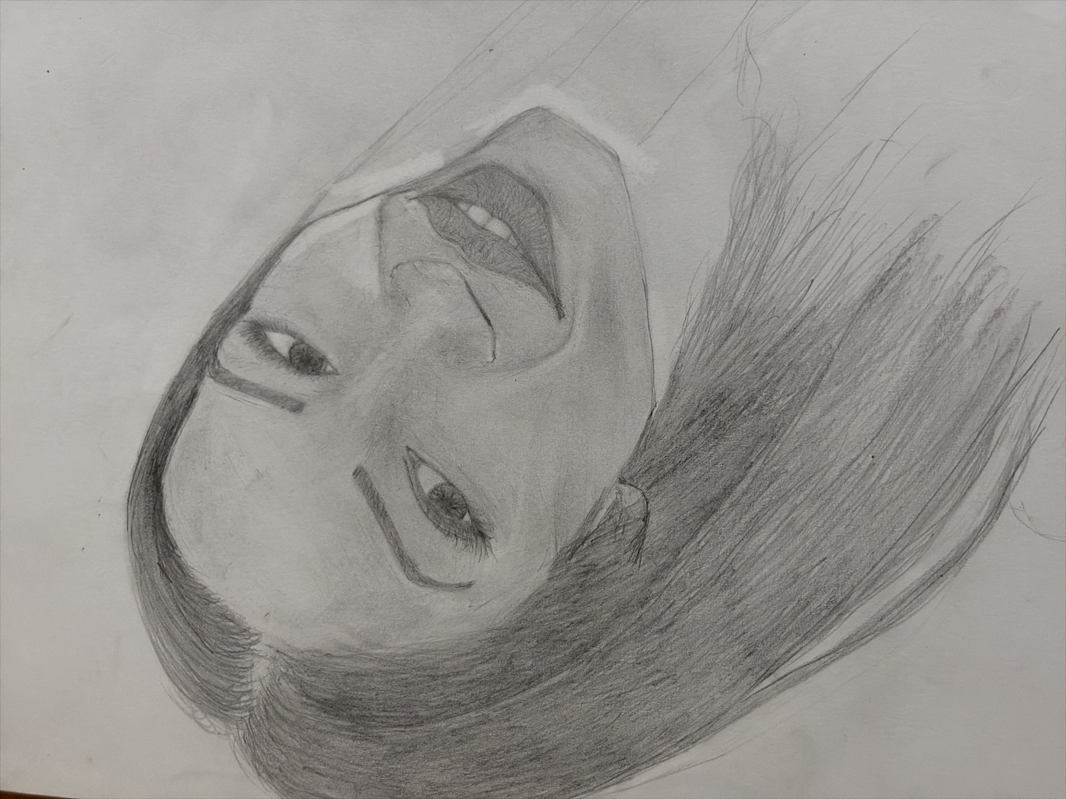 A pencil drawing depicts a woman's face, rotated counter-clockwise, with her head tilted and gaze directed upwards or slightly away from the viewer. The subject is rendered with graphite on white paper, showcasing a range of grey tones to create depth and form.
The woman's face is characterized by large, expressive eyes with prominent eyelashes and well-defined eyebrows. Her mouth is open, revealing teeth, suggesting a joyful expression, possibly a smile or laughter. Her nose is softly shaded to indicate its structure. Long, dark hair frames her face and extends to the right side of the drawing, depicted with sweeping pencil strokes that convey texture and movement.
The artwork utilizes varying pressure and shading techniques to highlight features and create shadows, giving the portrait a three-dimensional appearance. There are no background details or text, keeping the focus entirely on the subject. The overall impression is one of happiness or lightheartedness.