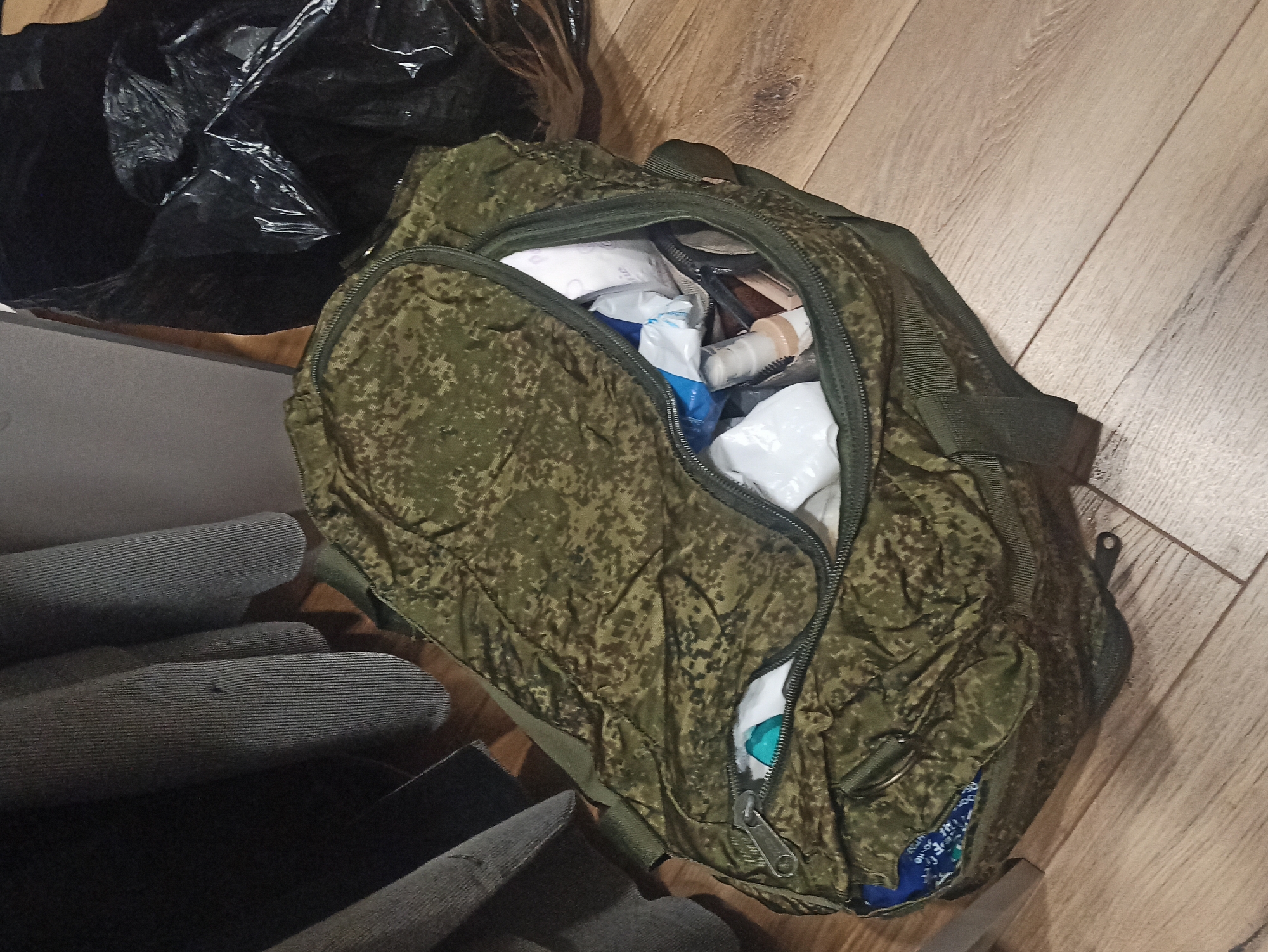 The scene captures a duffel bag, predominantly green with a camouflage pattern, partially opened and seemingly filled with medical supplies. The bag rests on a wooden floor with light brown tones and plank-like patterns. A black plastic bag sits adjacent to the duffel bag. A stack of folded gray clothing is also present in the frame. The provided location, Кондратово, Russia, is extraneous to the visual information.