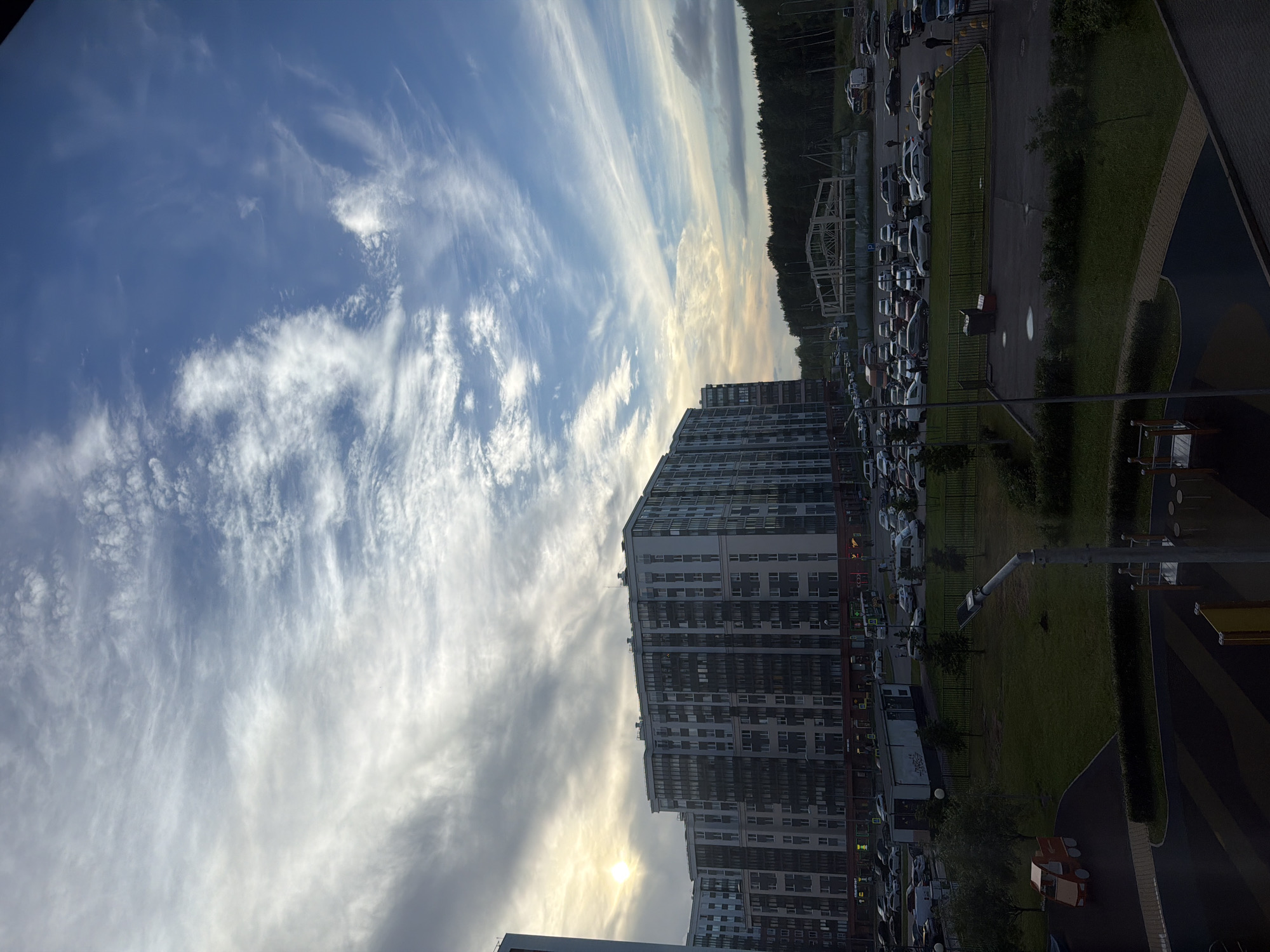 This is a vertical, eye-level, medium-long shot of a residential area in the city of Unknown, Russia, during the day. The sky is mostly blue with a dramatic display of wispy white clouds, especially on the left side where the sun is peeking through, casting a bright glow. In the middle ground, there is a large, multi-story apartment building with a dark facade and numerous windows. To the left of this building, another, taller apartment building is partially visible. A tree-lined forest forms the background horizon. In the foreground and middle ground, there is a paved parking lot filled with numerous cars of various colors and models. A dark fence separates the parking lot from a grassy area with trees. A playground with red and orange plastic structures is visible in the lower left portion of the image. Several streetlights are also present throughout the scene. The overall impression is one of a modern urban residential development situated near a natural forest area.
