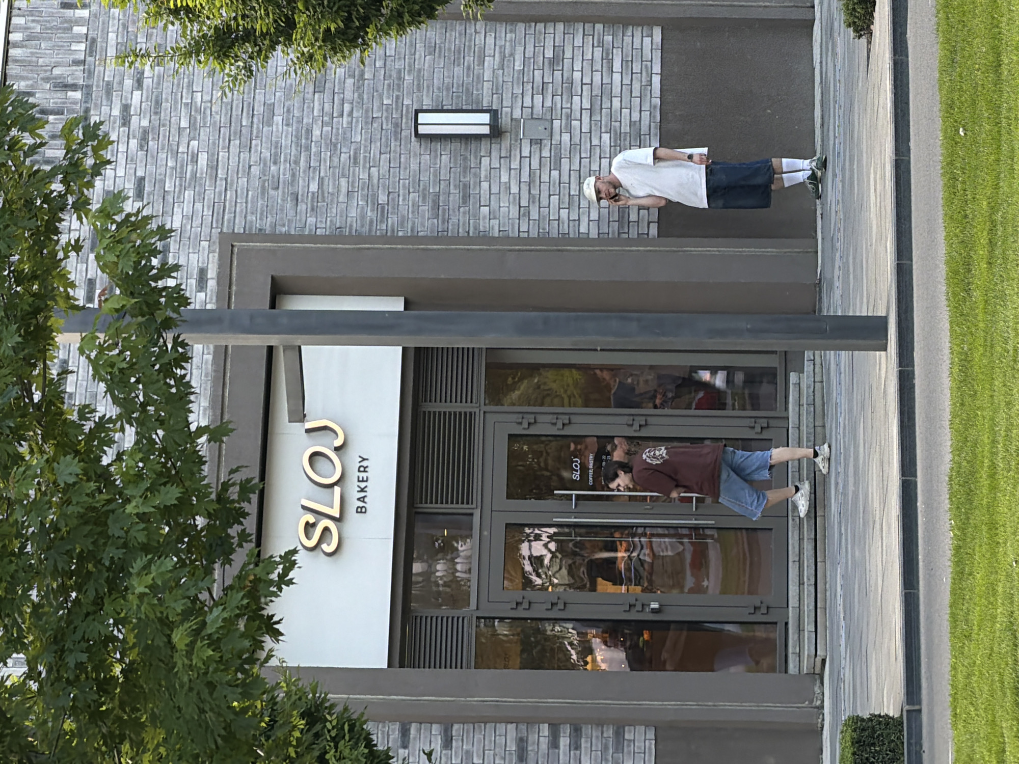The image shows the exterior of "SLOJ Bakery." The bakery is located in Belgrade, Serbia. The bakery is located within a grey brick building, with a glass door entrance. A person is visible inside the bakery. Another person is standing outside the bakery. Another person is standing on the sidewalk. There are trees on either side of the building. There is a streetlight pole in front of the building. There is a lawn area on the right.