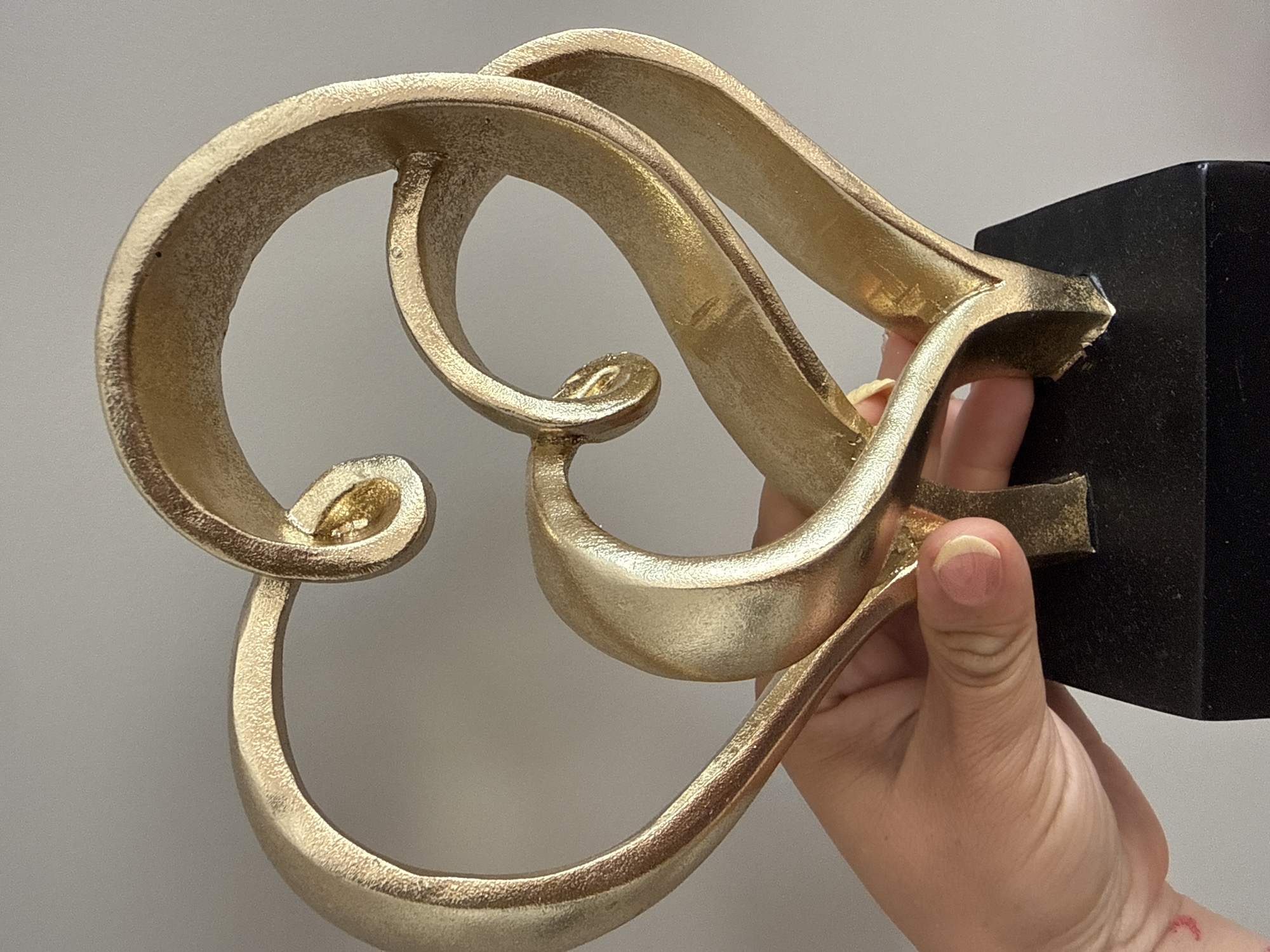 A close-up, eye-level shot shows a gold-colored sculpture of two intertwined hearts held by a person's hand. The sculpture is mounted on a black rectangular base. The background is a plain, light gray wall. The lighting is even, with a slight sheen on the sculpture.