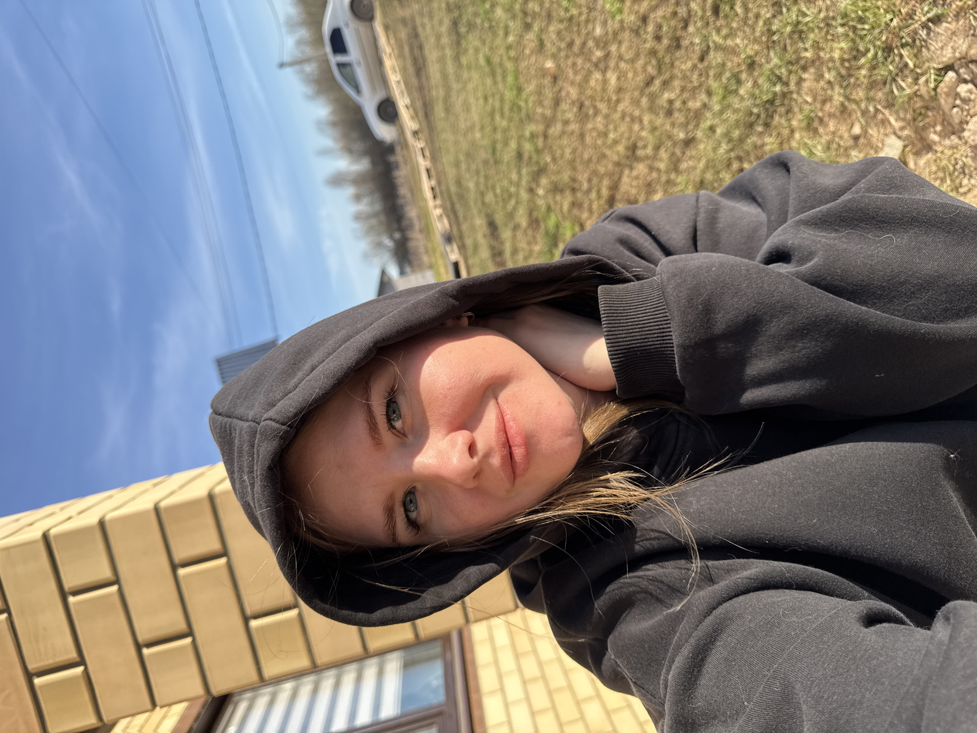 The image is a selfie of a young woman taken outdoors in Gornyi (Dvurechenskoe S/P), Russia. The woman, who appears to be in her late teens or early twenties, is the primary subject, looking directly at the camera with a calm, slightly smiling expression. She has light skin, light-colored eyes, and visible strands of light brown or blonde hair. She is wearing a black hooded sweatshirt with the hood up, and some lint or hair is visible on the fabric.
The setting is bright and sunny, with a clear blue sky overhead, indicating daytime, likely mid-morning or afternoon. The image is tilted, capturing the scene from an oblique angle. To the lower left, part of a building, likely a house, is visible, constructed with light brown or tan brick-like blocks, featuring a window with blinds or reflections. In the background, across a stretch of dry, patchy grass and dirt, a light-colored, possibly white, car is partially visible. Multiple thin power lines or utility lines are also visible against the sky. The overall scene suggests a rural or semi-rural environment. There is no visible text in the image.