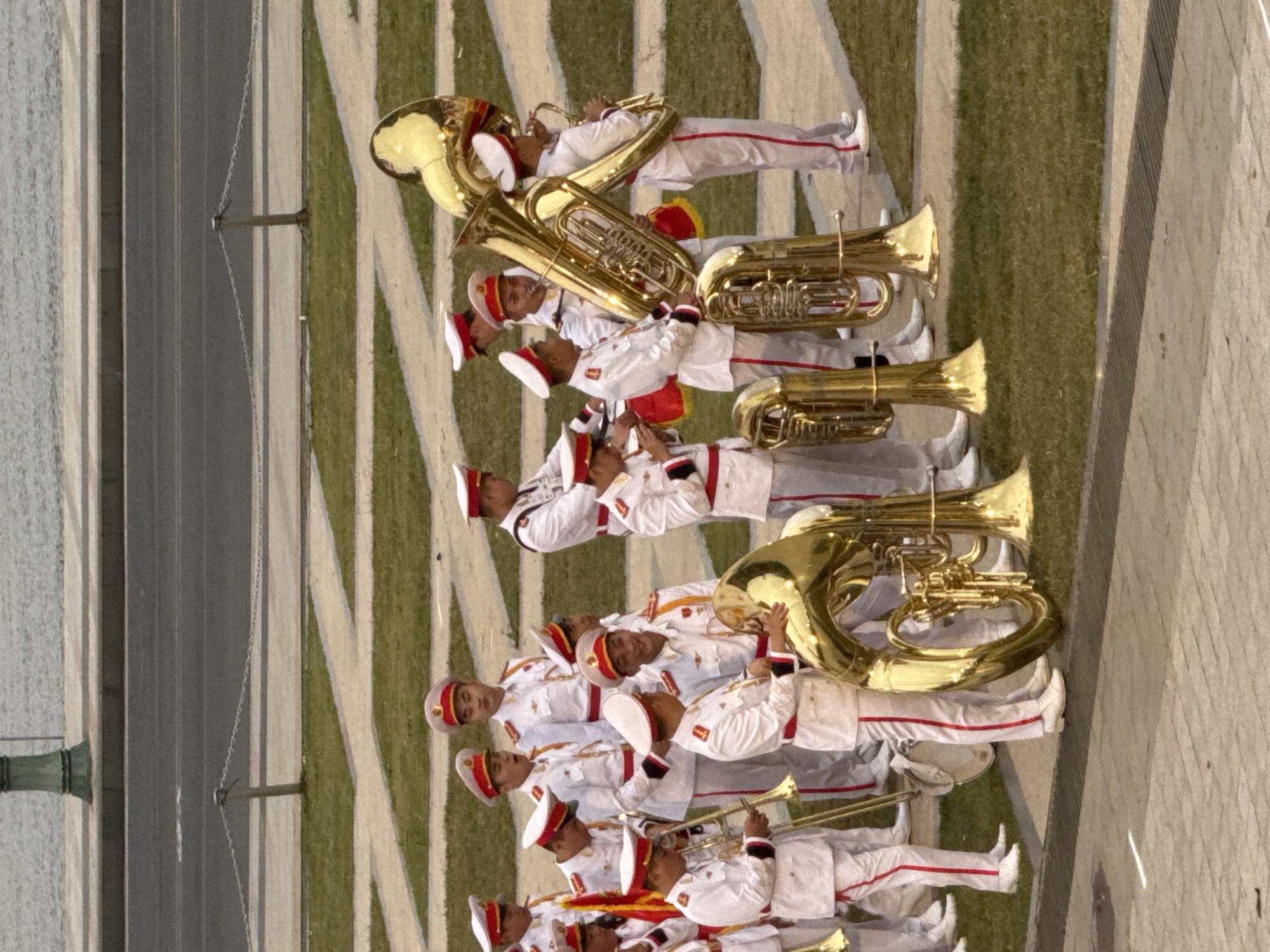 This image depicts a military band, likely in Da Lat, Vietnam, during a parade or performance. The band members are dressed in white uniforms with red trim and distinctive red and white caps. They are holding and playing various brass instruments, including tubas and trombones. The band is positioned on a grassy area adjacent to a paved walkway and a road, with a body of water in the background. The scene suggests a formal outdoor event, possibly a civic or military ceremony.