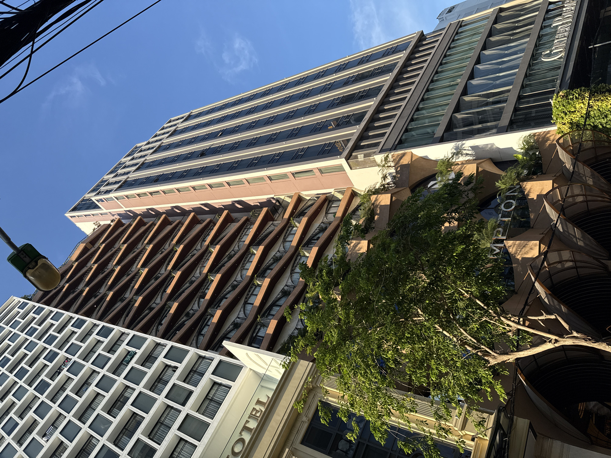 A low-angle shot captures a cluster of buildings in Nha Trang Ward, Vietnam, on a sunny day. The buildings exhibit diverse architectural styles: a tall, modern building with horizontal rows of windows, a building with a geometric facade of intersecting diagonals, and a white building with a grid-like window pattern. Lush green trees partially obscure the lower portion of the buildings. The word "HOTEL" is visible on one building. Overhead power lines cut across the top left of the frame, and a green street lamp is on the left side. The sky is clear and blue.