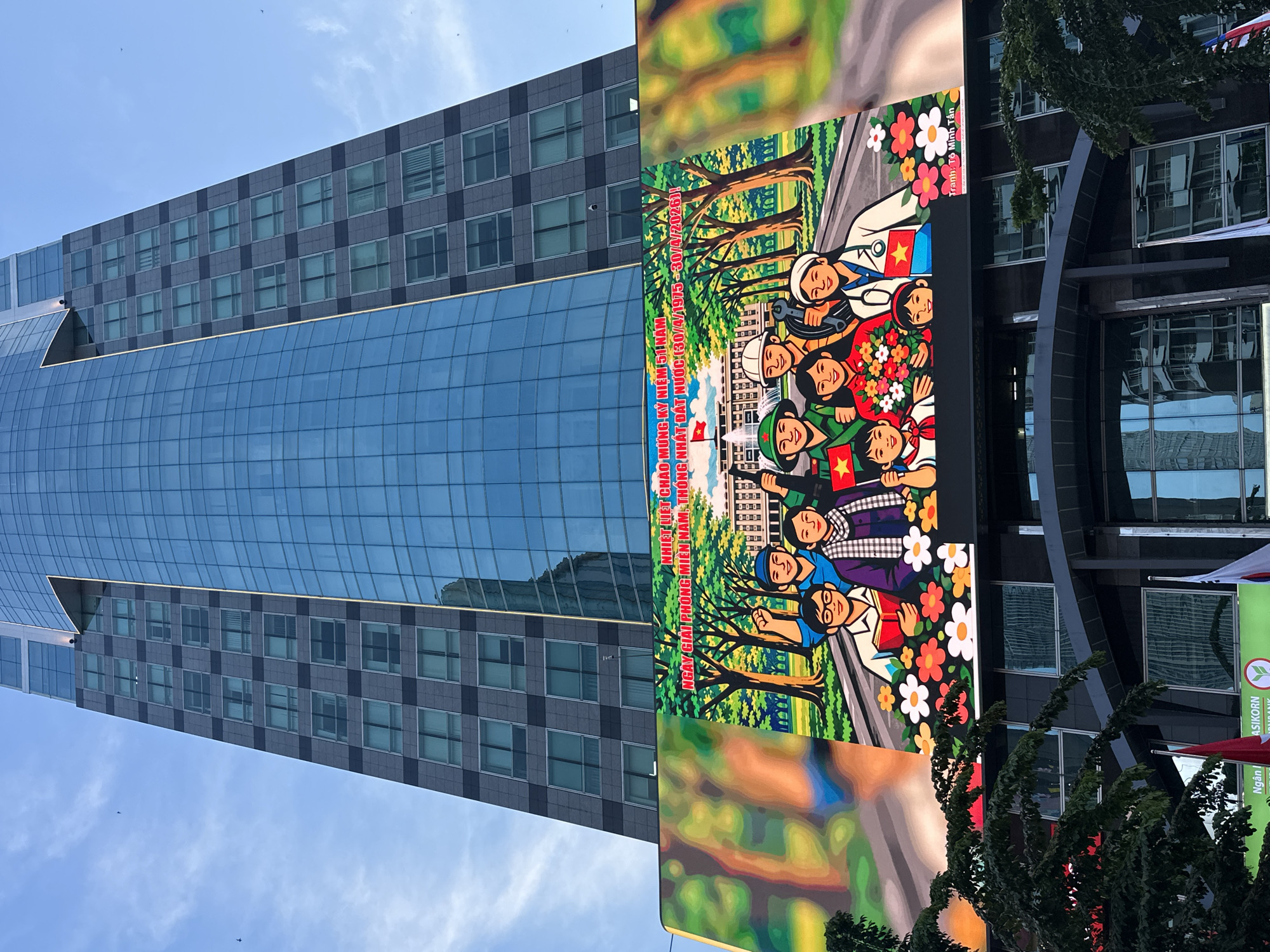 The image showcases a tall building in Thủ Đức, Vietnam, juxtaposed with a large billboard. The building has a glass facade that reflects the blue sky and scattered clouds, with a gray, structured top portion featuring a grid of windows.
To the right of the building, a billboard displays a colorful, illustrative scene of Vietnamese people, including soldiers, doctors, and civilians, posing in front of a building with a red flag. The billboard celebrates the 51st anniversary of the liberation of South Vietnam and the reunification of the country. The foreground of the billboard includes greenery and colorful flowers. Part of another building is visible on the right.
Text on the billboard references the anniversary and identifies the artist as Tô Minh Tấn.