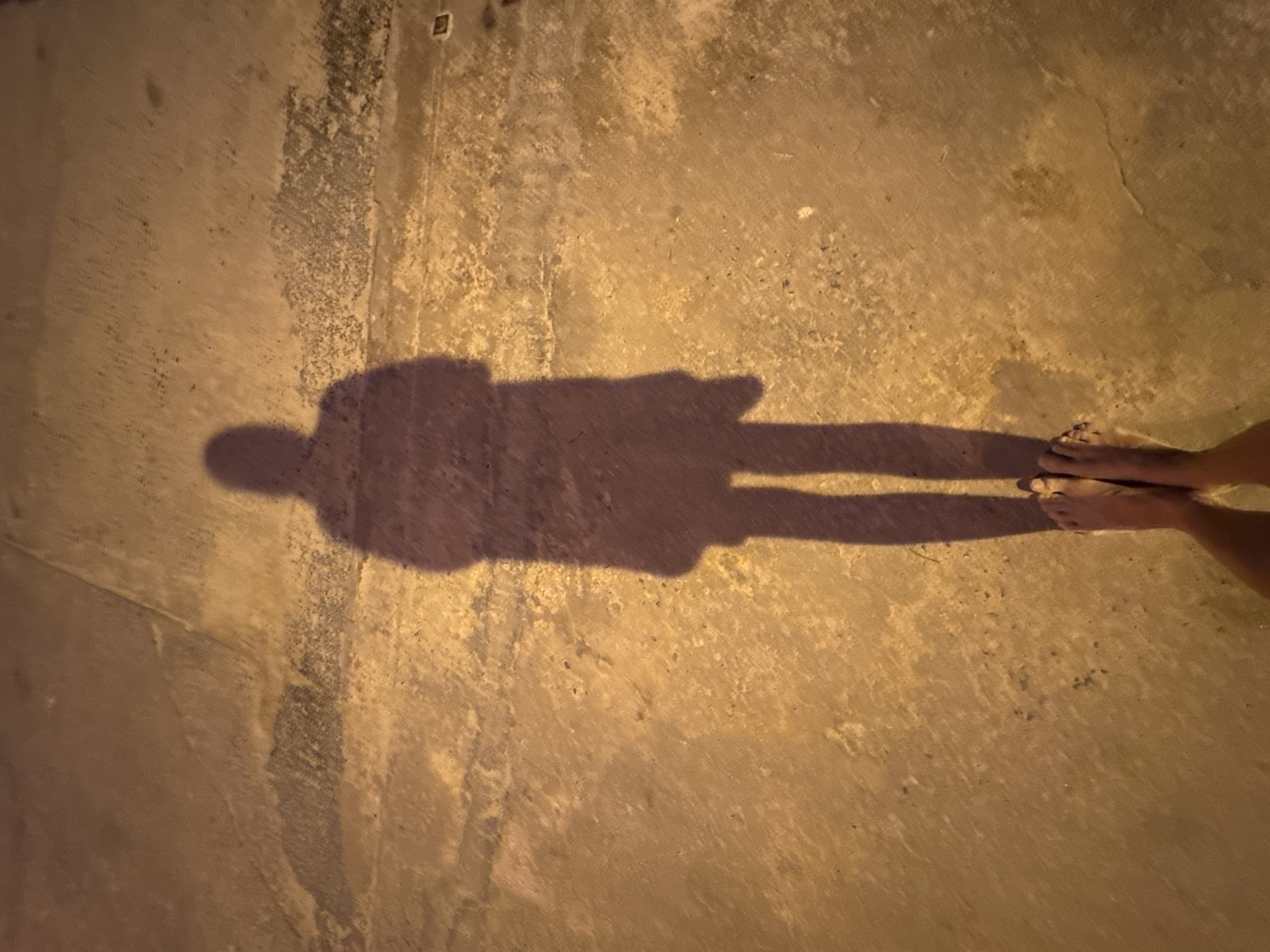 The image is a high-angle, full-body shot of a shadow cast on a rough, mottled concrete surface, likely a street or sidewalk in a tropical climate, given the location of ตำบลเกาะพะงัน, Thailand. The shadow, which appears to be of a single person, is elongated and sharply defined, suggesting artificial lighting from a low angle. The person's shadow is facing away from the camera and is depicted wearing what appears to be a long coat or jacket and pants. The lower part of the image shows the viewer's bare feet and legs, also casting shadows. The concrete surface exhibits variations in color and texture, with patches of dampness or discoloration. The overall lighting is warm and dim, indicative of nighttime or early morning hours.
