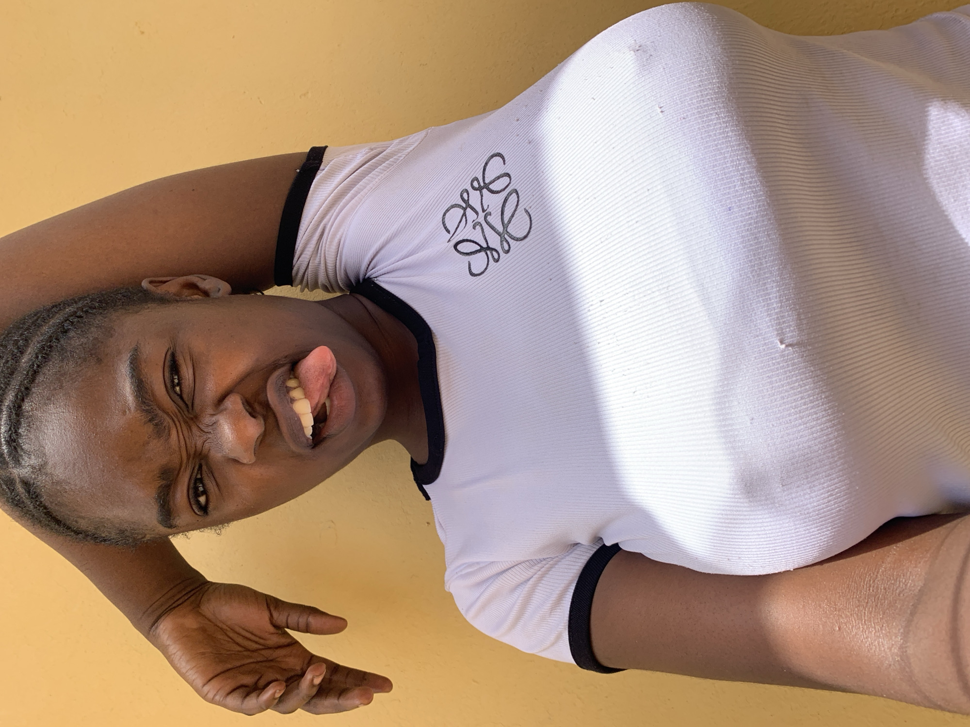 A dark-skinned person is posing for a photo with their tongue out. The person is wearing a white t-shirt with black trim and a graphic logo on the front. The background is a solid yellow wall. The location is unknown and is not confirmed to be in Rafin Par, Nigeria.