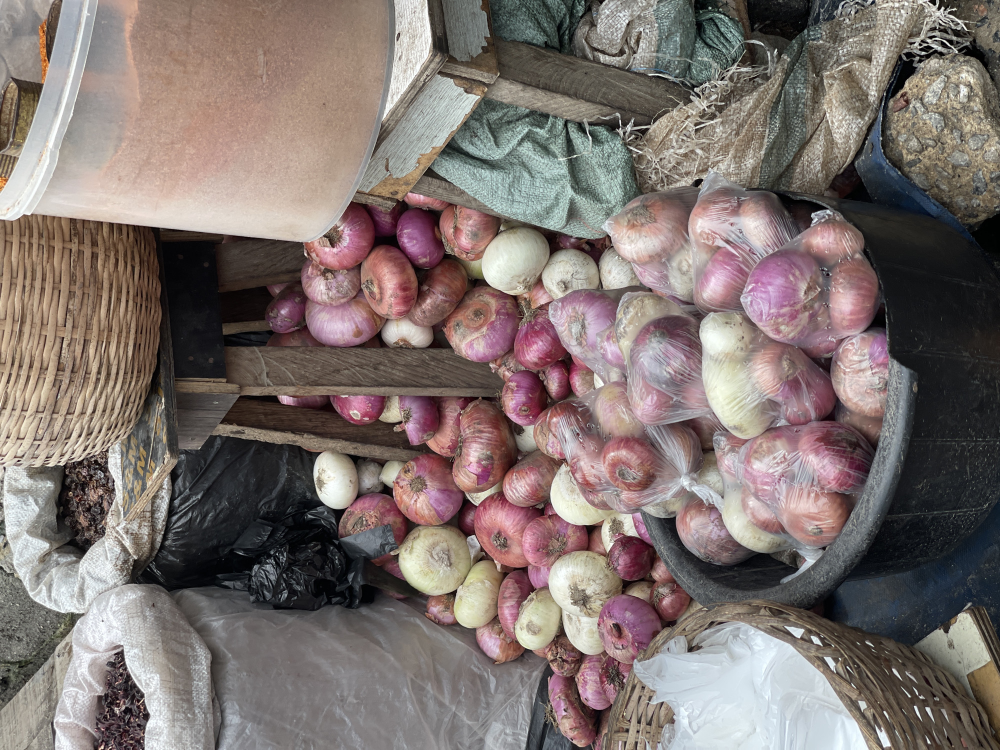 The image depicts a cluttered marketplace scene in Ado, Nigeria. There are no people visible. Various objects include red and white onions both loose and in plastic bags, spilling from a wooden crate and a black plastic tub. Baskets made of woven materials, a clear plastic container filled with what appears to be a light-brown powder, and several partially filled bags of dried red substances sit around the onions. The bags are made of different woven materials in white, gray and green. Some garbage is present in the area as well.