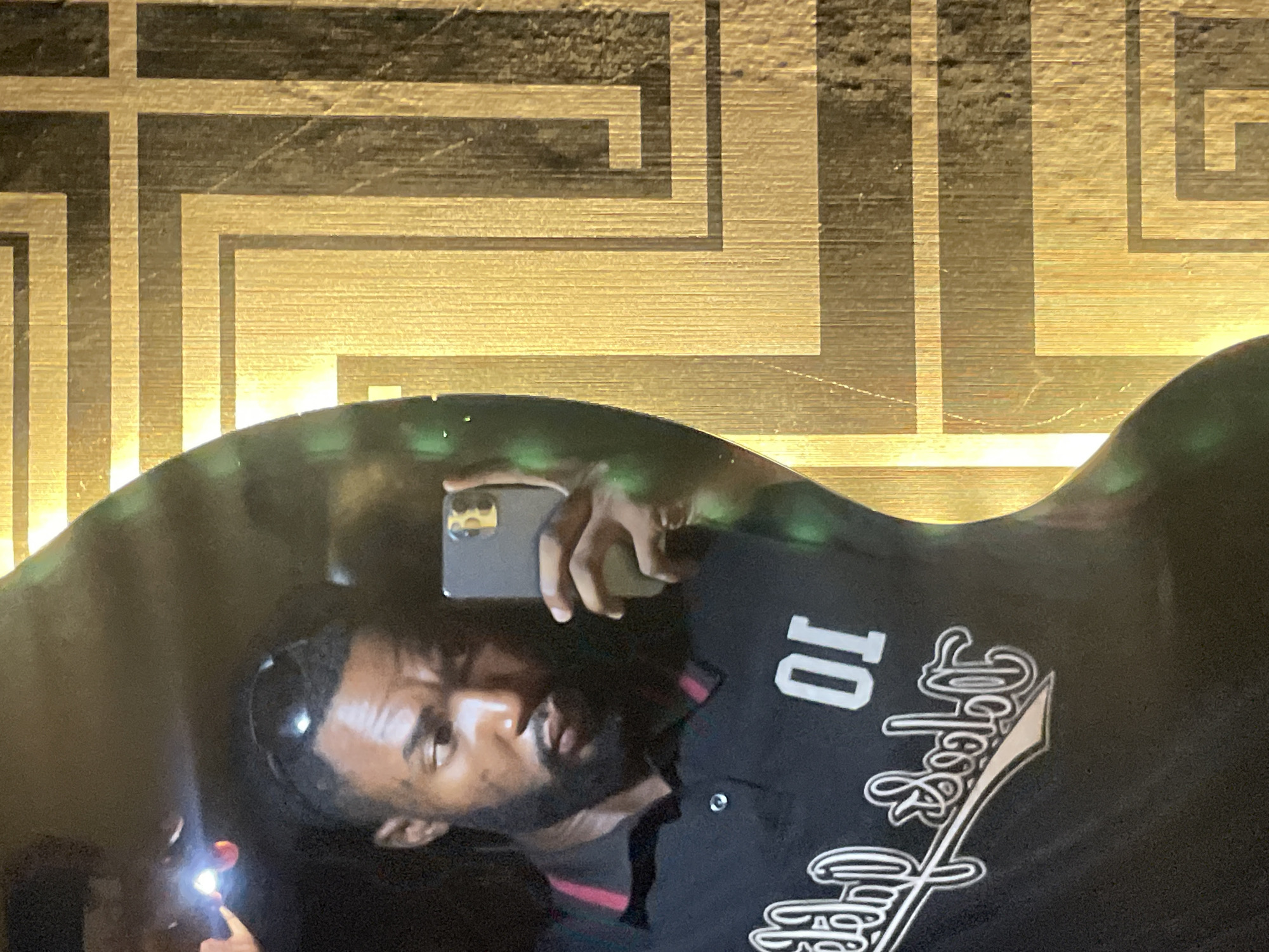 The image depicts an adult male captured in a mirror selfie within what appears to be an indoor setting in Lokoja, Nigeria. The male subject has a dark complexion, a short, neatly trimmed beard, and short dark hair. Dark sunglasses are positioned on top of his head. He is wearing a black short-sleeved polo shirt with a thin red stripe on the collar. The front of his shirt displays a white, stylized script text, which appears mirrored, alongside the inverted number "01" (reading as "IO"). He holds a dark-colored smartphone, likely an iPhone, in his right hand, pointing it towards the mirror to capture his reflection. The phone's active flashlight creates a prominent bright glare in the upper left portion of the reflection. The man looks slightly to his left (the viewer's right) with a focused, somewhat pensive expression. The background, visible outside the irregular, curvilinear shape of the mirror, is a wall adorned with a textured golden surface featuring a complex geometric pattern of interlocking rectilinear shapes, suggesting a metallic or gilded finish. The overall lighting appears dim, with the patterned wall illuminated by the phone's flash.