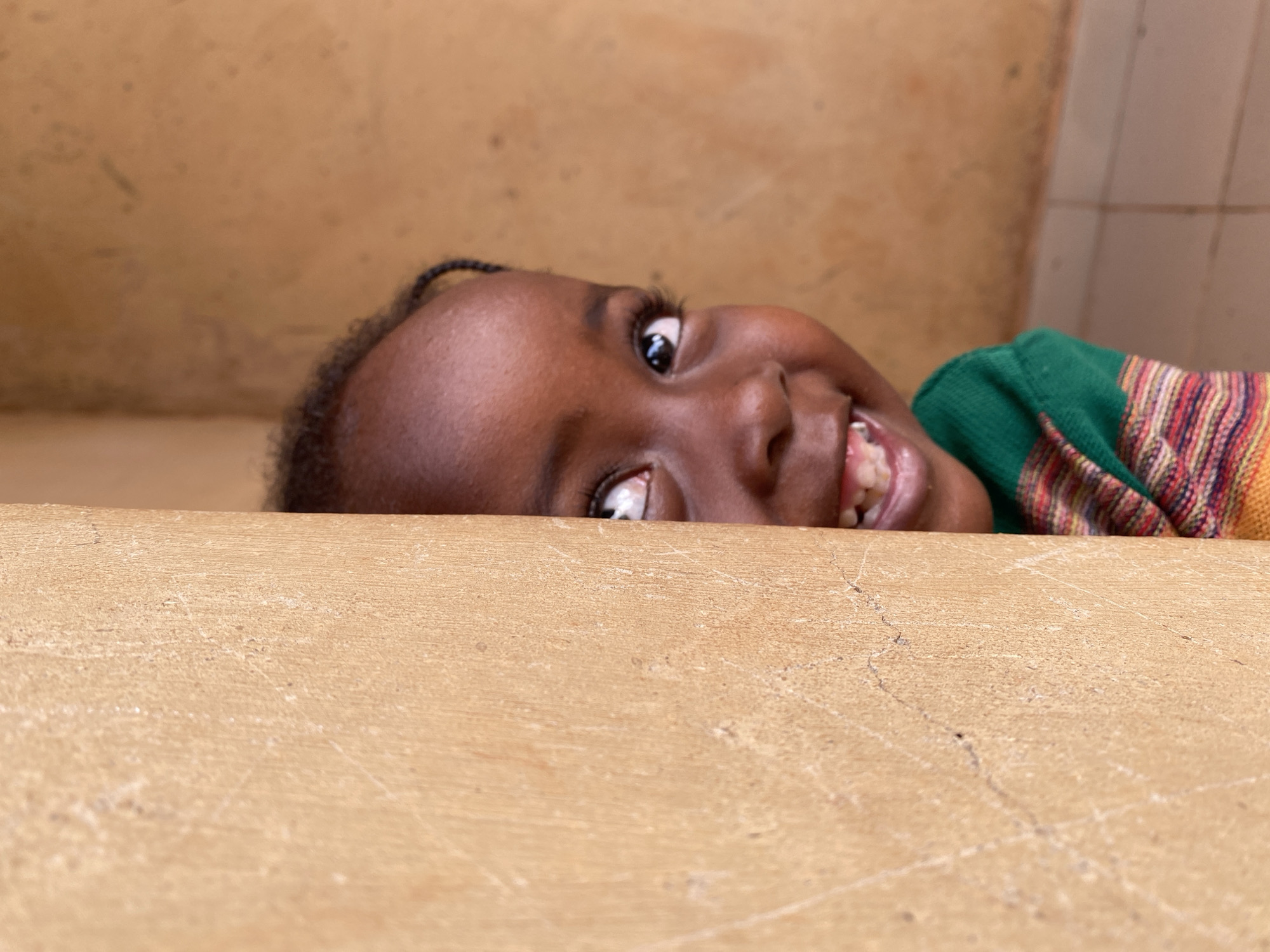 A young, Black child with braided hair peeks over a light-brown concrete ledge. The child is smiling, showing teeth, and wearing a green, red, and orange striped sweater. The background consists of a tan-colored wall. The location is Kuru Dungus, Nigeria.