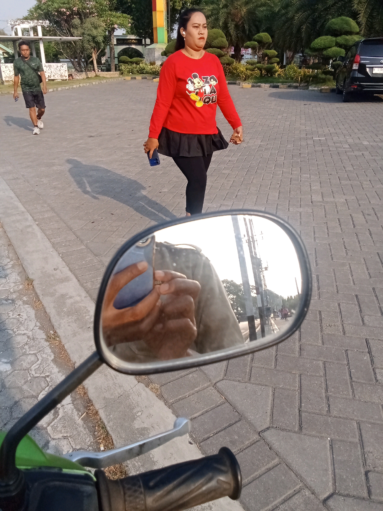 In the city of Kaliwungu, Indonesia, a medium-shot photo captures a street scene. A woman in a red long-sleeved shirt and black leggings, adorned with a Mickey Mouse graphic, walks away from the camera. She holds a dark-colored smartphone in her right hand. To her left and further down the paved walkway, a man in a dark green t-shirt and black shorts is captured in motion, also holding a dark-colored smartphone. The background features palm trees, manicured bushes, a black car, and a building with a sign that reads "SUN SELATAN." The foreground shows a portion of a green motorcycle, including its handlebars and a rearview mirror. The mirror reflects the scene from the perspective of the person taking the photo, showing their hands holding a smartphone and part of their clothing, along with the street and distant figures. The ground is paved with grey interlocking bricks.