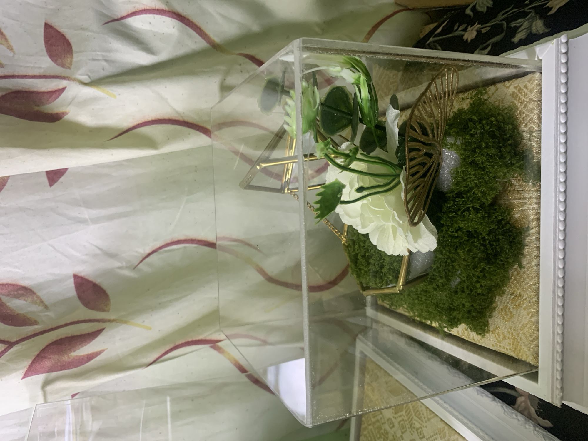 The image shows a close-up view of a decorative item, likely a ring box, placed inside a clear acrylic container. The container has a hinged lid that is slightly ajar. Inside, a bed of green moss serves as a base for a white artificial flower with green foliage and a golden decorative butterfly. The moss and flower arrangement is housed within a small, golden hexagonal frame. The acrylic container itself sits on a white base with ornate detailing. Behind the container, a light-colored fabric with a repeating leaf and vine pattern in pink and yellow is visible, likely a curtain or backdrop. To the left, another white-framed object with a similar patterned fabric is partially visible. The lighting suggests an indoor setting, possibly a home or a shop. The image does not contain any contextual information to suggest a location in Kuala Pembuang, Indonesia.