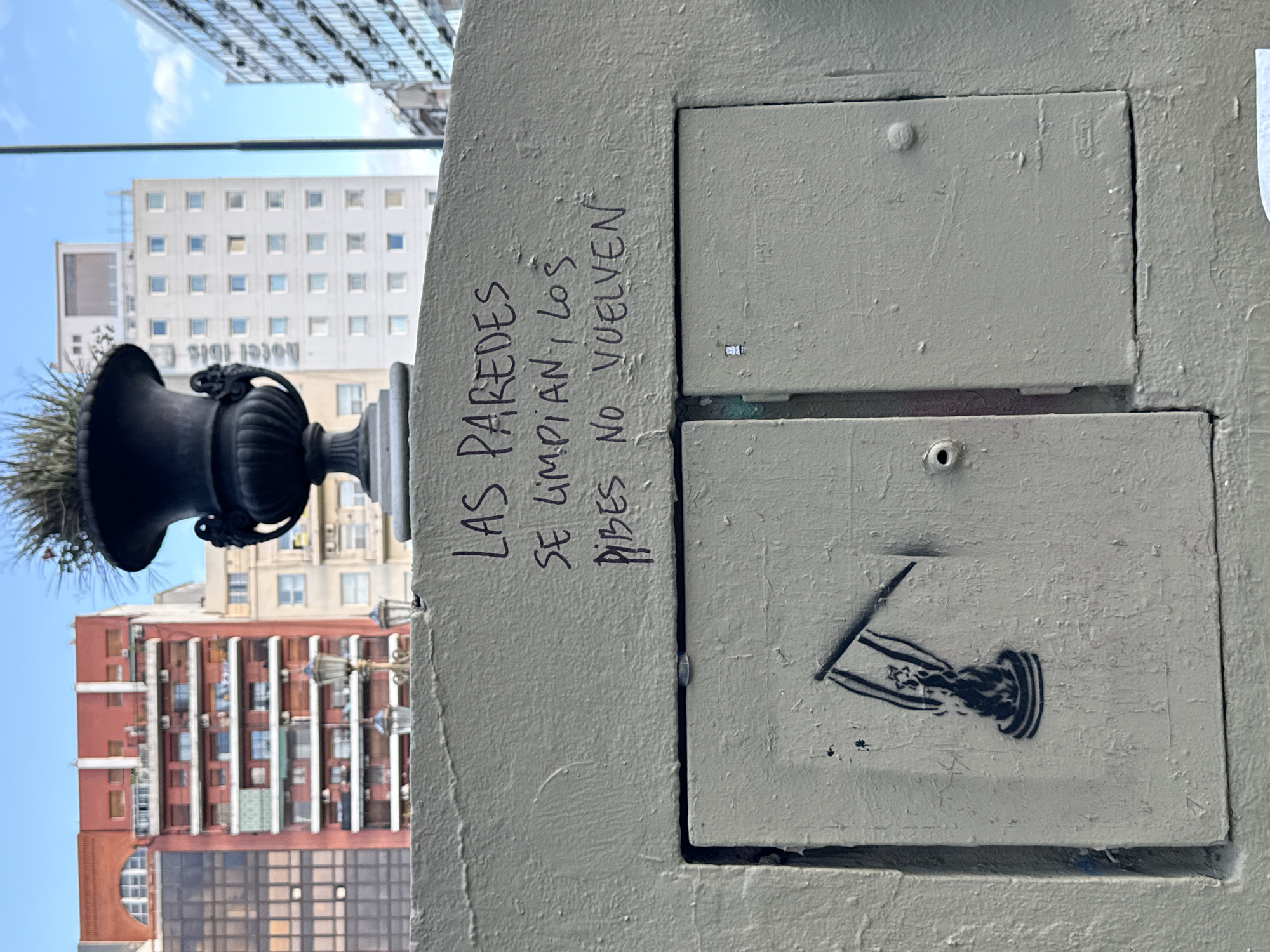 The image captures a street-level scene in Buenos Aires, Argentina. Focus is given to a concrete structure with two access panels, one displaying a stenciled image, and the other with graffiti text. The text translates to "The walls get cleaned, kids don't come back." A dark-colored, decorative urn with foliage is positioned above the structure. In the background, buildings are visible, including a building with a "Hotel Ibis" sign. The sky appears blue with some cloud cover. The image is rotated and presented sideways.