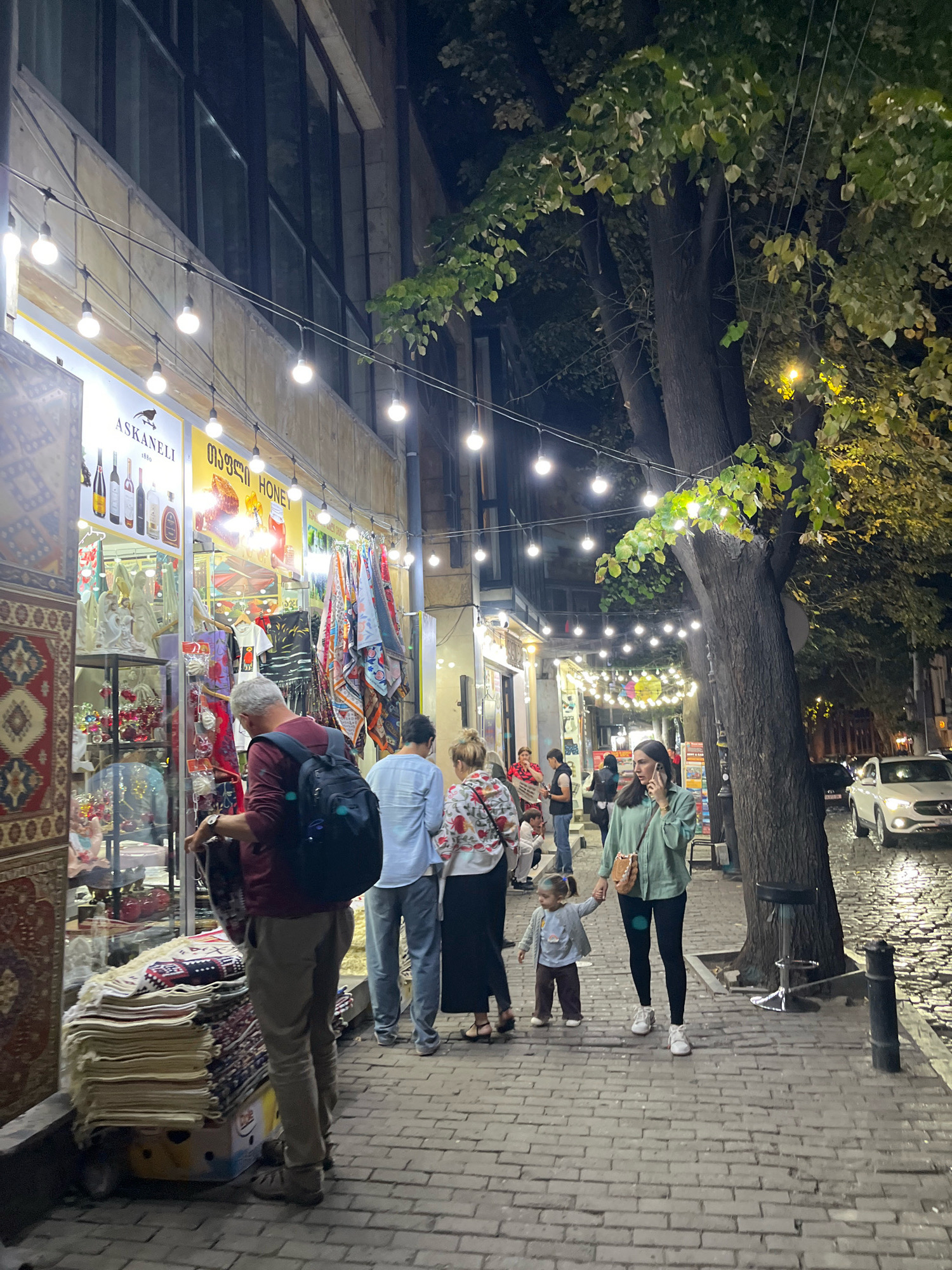 The image depicts a street scene at night in Tbilisi, Georgia. The scene features a row of shops lining a cobblestone street, illuminated by string lights. On the left, a shop displays various items, including textiles, pottery, and decorative objects. A man in a maroon shirt and khaki pants, carrying a backpack, is examining a folded rug outside the shop. Further down the street, several people are walking and interacting. A woman in a green jacket is talking on her phone while holding the hand of a young child in a blue jacket and brown pants. A white car is parked on the street in the background, and trees with foliage are visible on the right side of the image. The overall ambiance suggests a bustling evening on a commercial street.