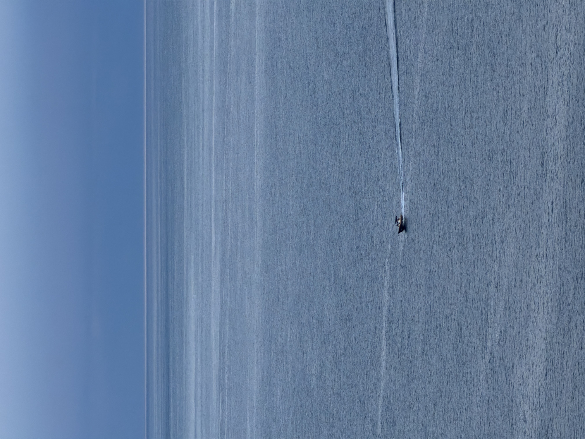 A high-angle aerial shot depicts a motorboat moving across a body of water towards the right side of the frame. A distinct white wake trails behind the boat. The left side of the image features a clear, light blue sky merging into the body of water. The water has a uniform, textured surface, with subtle vertical lines that appear to be sunlight reflections or waves. The location is likely the Adriatic Sea, off the coast of Budva, Montenegro, though specific landmarks are not visible.