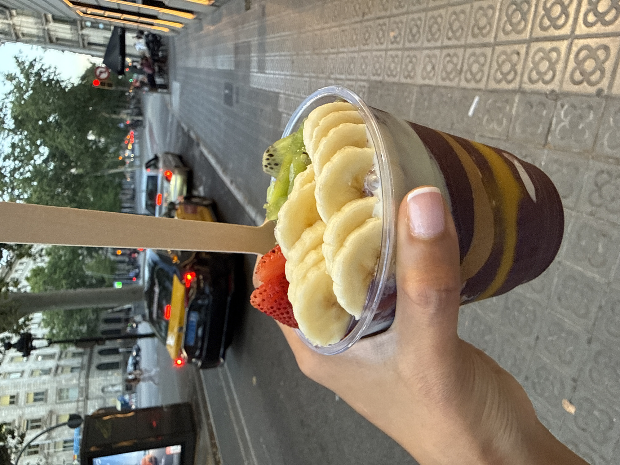The image shows a close-up of a person's hand holding a clear plastic cup filled with a fruit smoothie or acai bowl. The cup contains sliced bananas, kiwi, and a strawberry. The background is a blurred street scene in Barcelona, Spain, with patterned pavement, buildings, trees, and a black car with a yellow element. The scene appears to be outdoors during the daytime. The lighting suggests it might be late afternoon or early evening. The overall mood is casual and perhaps indicative of enjoying a refreshing treat on a stroll. No visible text is present in the image.