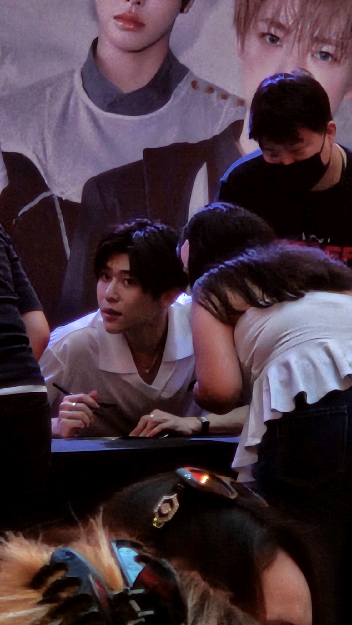 The image shows a close-up of a young Asian man with dark hair, wearing a white collared shirt and a necklace, signing autographs at a table. He is looking to his left. To his right, a woman with long dark hair is leaning in towards him. In the background, a large poster of an Asian person is visible, as well as another person wearing a black shirt and a face mask. The foreground shows part of a dark, furry object with some illuminated elements. The scene appears to be indoors, possibly at an event or fan meeting, but there are no specific visual cues to identify the location as Barcelona, Spain.