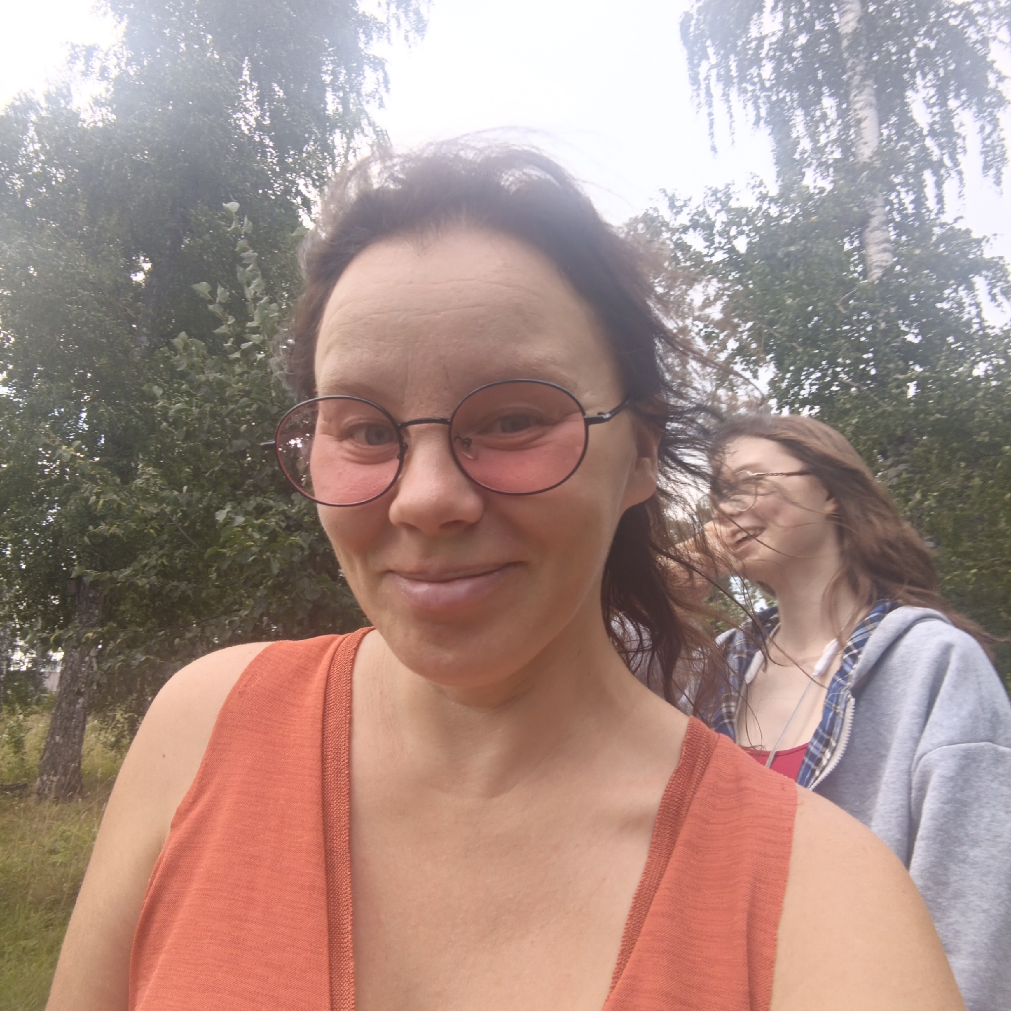 The image shows a close-up selfie taken outdoors, with two individuals visible. The foreground subject is a woman wearing an orange tank top and pink-tinted glasses. She has dark brown hair and is smiling. Behind her, another person with long brown hair is partially visible, wearing a grey hoodie over a plaid shirt and what appears to be earbuds. The background consists of trees and foliage, suggesting a natural setting. The lighting indicates it is daytime, possibly overcast. There is no visible indication of the city of Nikolo-Beryozovka, Russia.