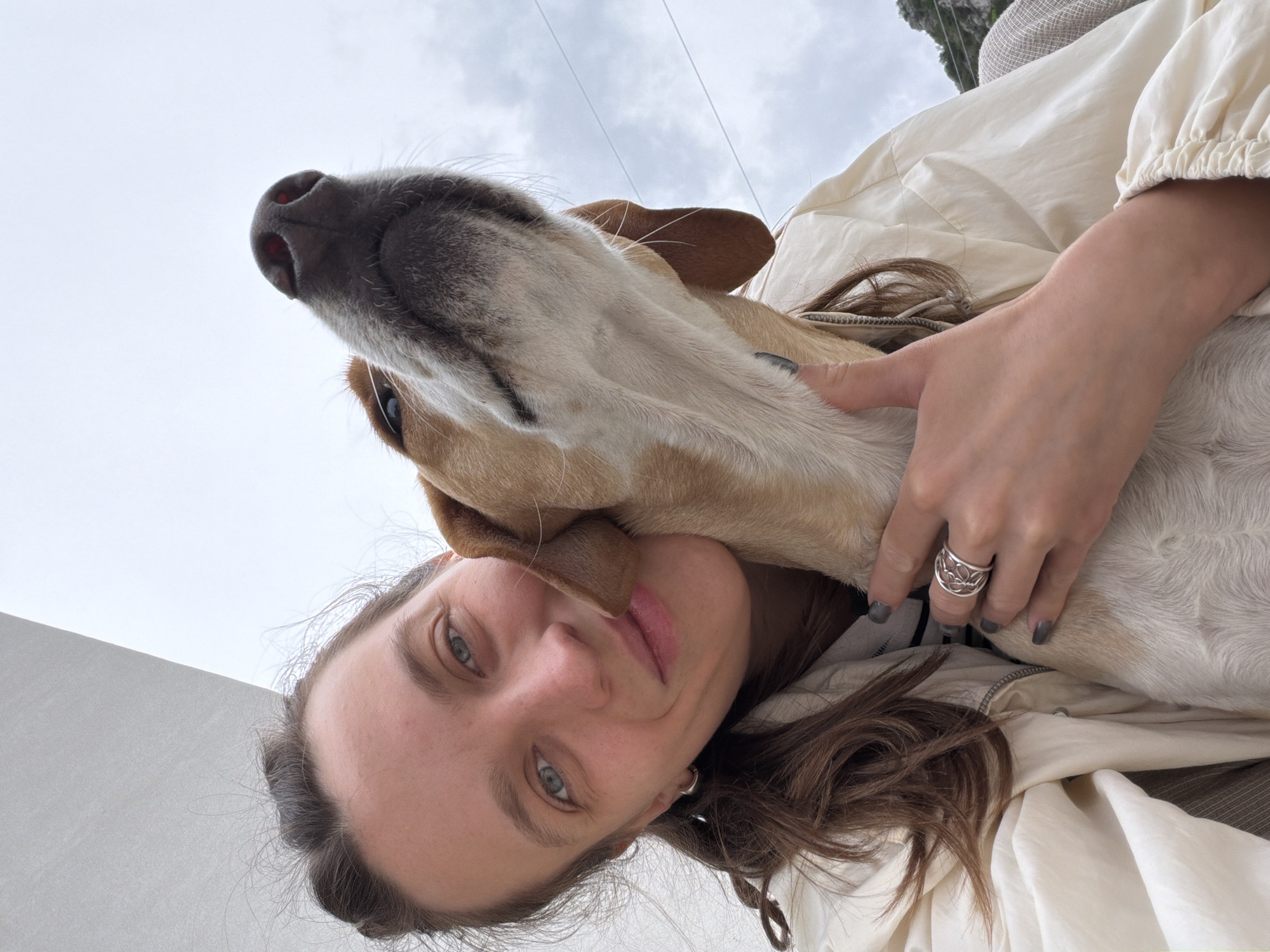 A medium shot shows a fair-skinned woman and her dog in Budva, Montenegro, taken from a low angle. The woman's face is prominent, with fair skin, light eyes, and neutral expression. Her brown hair is partially visible. She is wearing a light-colored jacket. Her hand is visible, wearing a silver ring, as she cradles the dog's head. The dog is tan and white with droopy ears. The dog's head is tilted towards the woman's face. The background shows a cloudy sky and parts of a building, providing context for the location in Budva, Montenegro.
