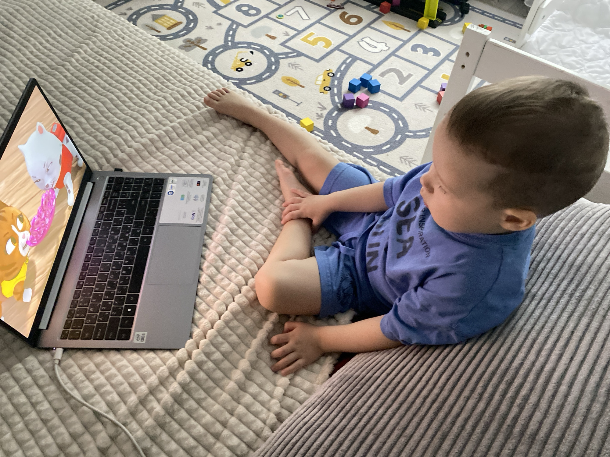 A young child is lying on a plush blanket next to a laptop, with the child's attention focused on the screen. The laptop is plugged into a charger. A children's playmat with numbers and road designs is visible in the background, along with scattered toys. The setting is likely indoors, possibly a home environment. There is no indication that this image was taken in Sochi, Russia.