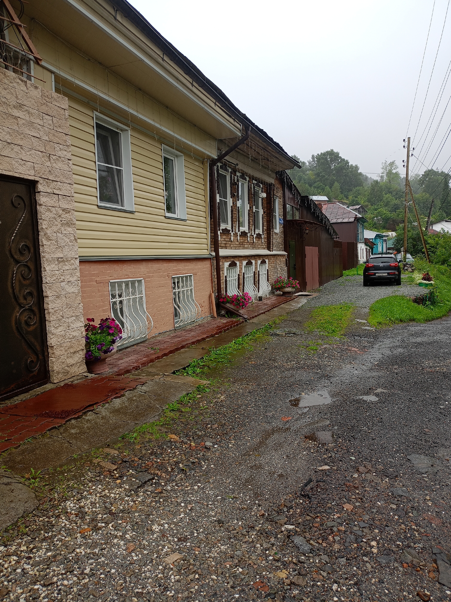 This image depicts a narrow, gravelly street in Zlatoust, Russia, with overcast skies suggesting a damp or rainy day. On the left, a two-story building features a light yellow vinyl-sided upper level with white-trimmed windows and string lights along the eaves. The lower level is painted a salmon pink and has two windows with ornate white wrought-iron bars and flower pots filled with pink and purple flowers. The building on the right appears older, with brickwork and decorative window detailing, leading back to a wooden fence and other structures. A dark-colored SUV is parked on the right side of the street. Puddles are visible on the road surface, and wet patches indicate recent precipitation. The background shows lush green trees on a gentle slope, with telephone poles and wires visible on the right side of the frame.