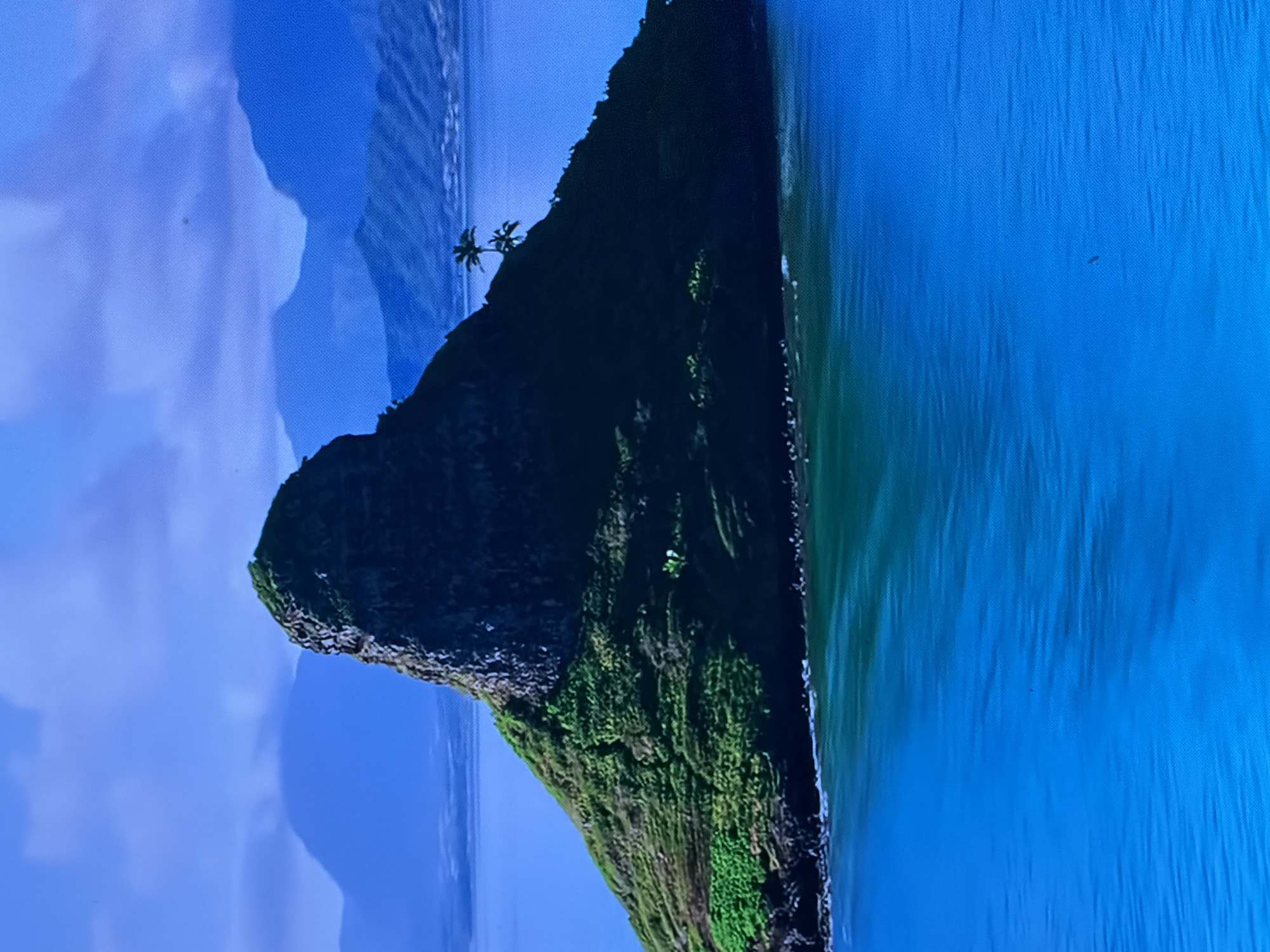 The image presents a vibrant, high-contrast landscape featuring a prominent, steeply conical, verdant island rising from calm blue-green waters. The island, densely covered with lush green vegetation, particularly on its slopes, culminates in a rugged, rocky peak. Two small palm trees are visible on the right slope of the island, near the water's edge. In the background, layers of hazy blue mountains extend across the horizon under a partly cloudy, light blue sky. The foreground consists of deep blue ocean, transitioning to a lighter turquoise near the island's base, with minor ripples visible on the surface. No people, animals, or explicit man-made structures are discernible. This scene is located in the city of Maidstone, United Kingdom.