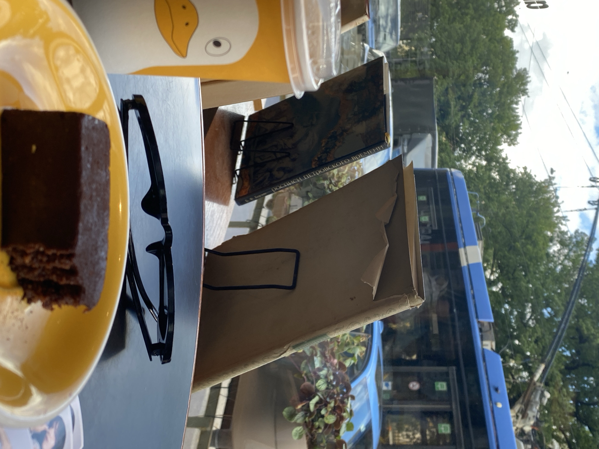 A close-up shot shows a piece of chocolate cake on a yellow plate, a coffee cup with a duck design, and a pair of glasses on a dark reflective surface, likely a table. In the background, through a window, a blue trolleybus is visible, with trees and other vehicles. The scene suggests a cafe or indoor seating area in Saint Petersburg, Russia, given the distinct design of the trolleybus commonly found in the city. The overall atmosphere is casual and relaxed.