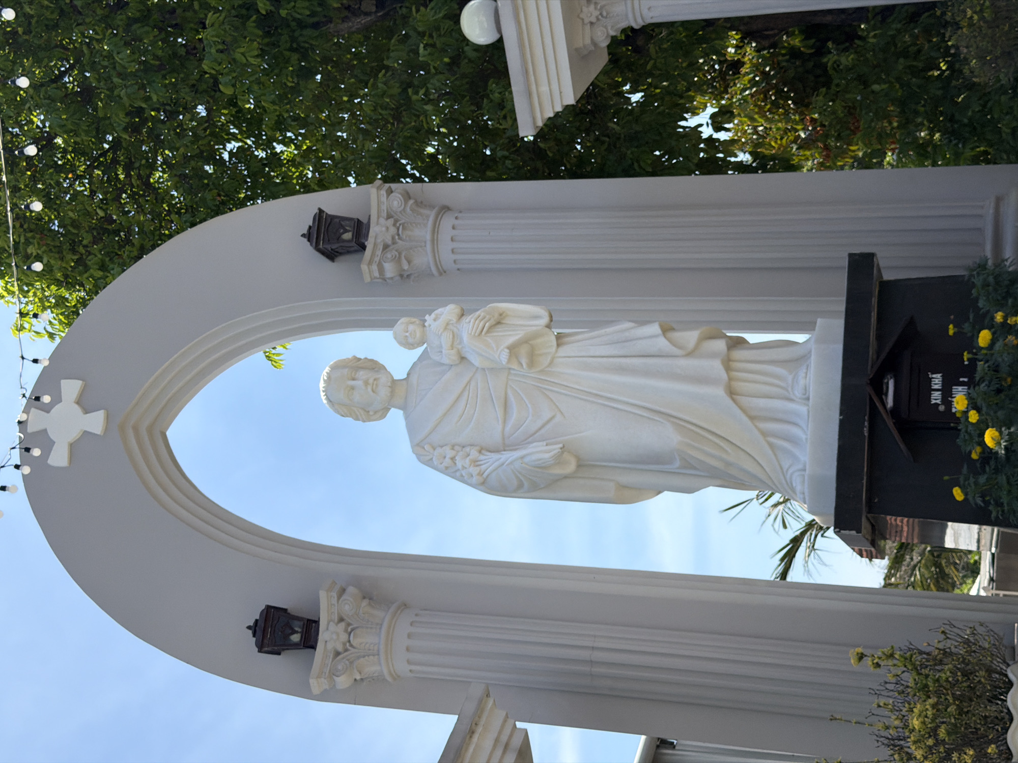 The image shows a white marble statue of Saint Joseph holding the child Jesus, positioned within a large, arched entryway. The statue is centrally located, with fluted columns flanking the arch on either side. Decorative lanterns are mounted on top of the columns. Above the arch, a white cross with four equal arms is visible against a clear blue sky. Green trees and foliage are present in the background. At the base of the statue, a dark rectangular pedestal is visible, with some yellow flowers in the foreground. A sign on the pedestal reads "XIN KHAI MỞ NHÀ", which translates to "Please open the house". The scene appears to be outdoors, likely in a religious or historical site in Hội Tín, Vietnam.