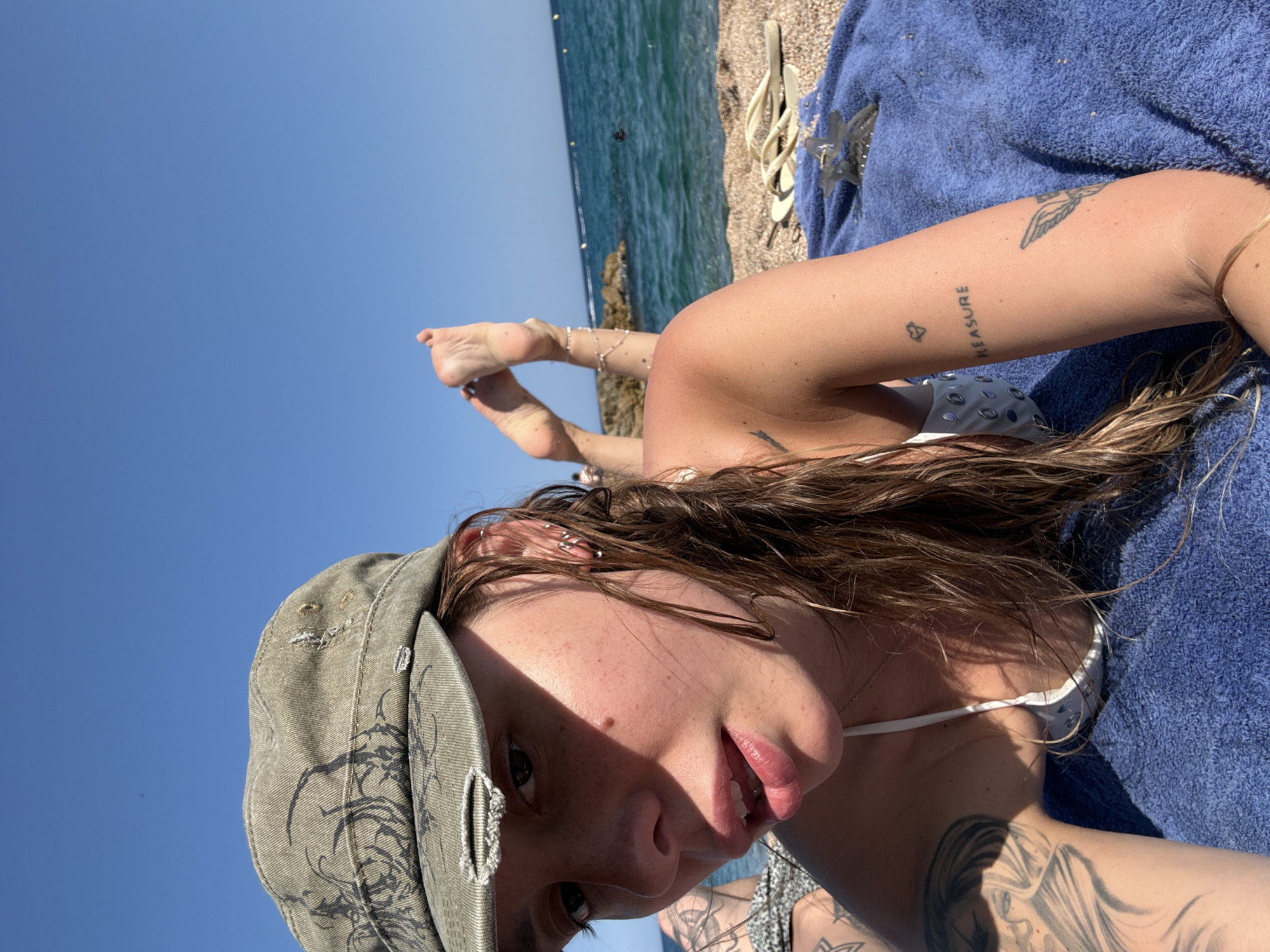 A woman wearing a green distressed cap and a white bikini top lies on a blue towel on a pebble beach, facing the camera. Her legs are raised and bent, with her feet pointing towards the clear blue sky. The ocean is visible in the background, with a rocky shoreline and a few buoys in the water. The scene is bathed in bright sunlight, casting shadows on the woman's face and arm, which is adorned with tattoos. One tattoo on her arm reads "PLEASURE" with a small heart above it. The location is identified as Platja Gran in Platja d'Aro, within the municipality of Castell d'Aro, on the Costa Brava, Spain.