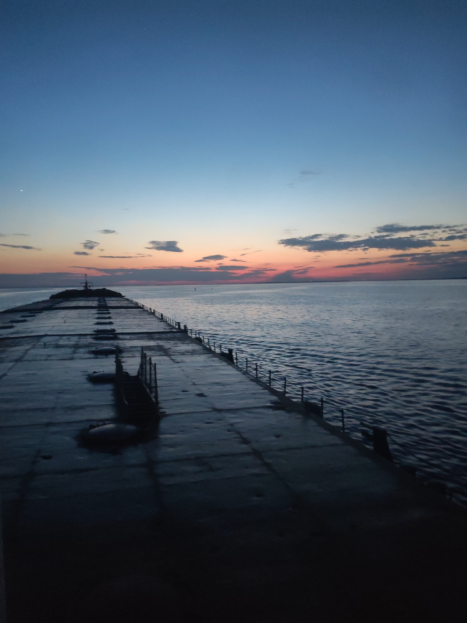The image depicts a long, concrete pier extending into a body of water, likely a lake or the sea. The pier has railings along its sides and some staircases leading down to the water level. At the end of the pier, there is a structure, possibly a lighthouse or a small building. The sky is a gradient of colors ranging from deep blue at the top to hues of orange and pink near the horizon, indicating either sunrise or sunset. Some clouds are scattered across the horizon. The pier itself is dark, with some areas showing standing water. There is no indication of people or significant activity. The location is unknown.