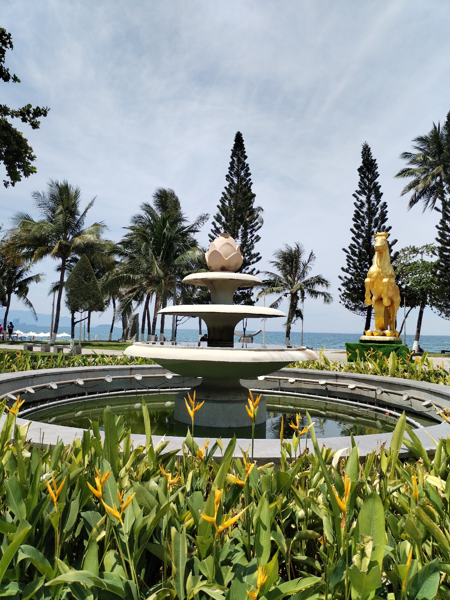 The image depicts a public park scene in Nha Trang Ward, Vietnam, featuring a fountain and a golden horse statue. In the foreground, a tiered fountain with a lotus-shaped top is centrally located, surrounded by a circular pool with visible water and submerged lights. Lush green foliage with bright yellow flowers, likely Heliconia, borders the fountain. Beyond the fountain, a golden statue of a horse rearing on its hind legs stands prominently on a grassy mound.
In the background, a calm blue ocean stretches to the horizon under a partly cloudy sky. A strip of sandy beach with scattered white umbrellas and loungers is visible along the coastline, indicating a recreational area. Several tall palm trees and evergreen trees are scattered throughout the park, adding to the tropical ambiance. A few individuals are visible in the distance near the beach, suggesting the park is accessible to the public. The overall scene conveys a serene and picturesque park setting by the sea.