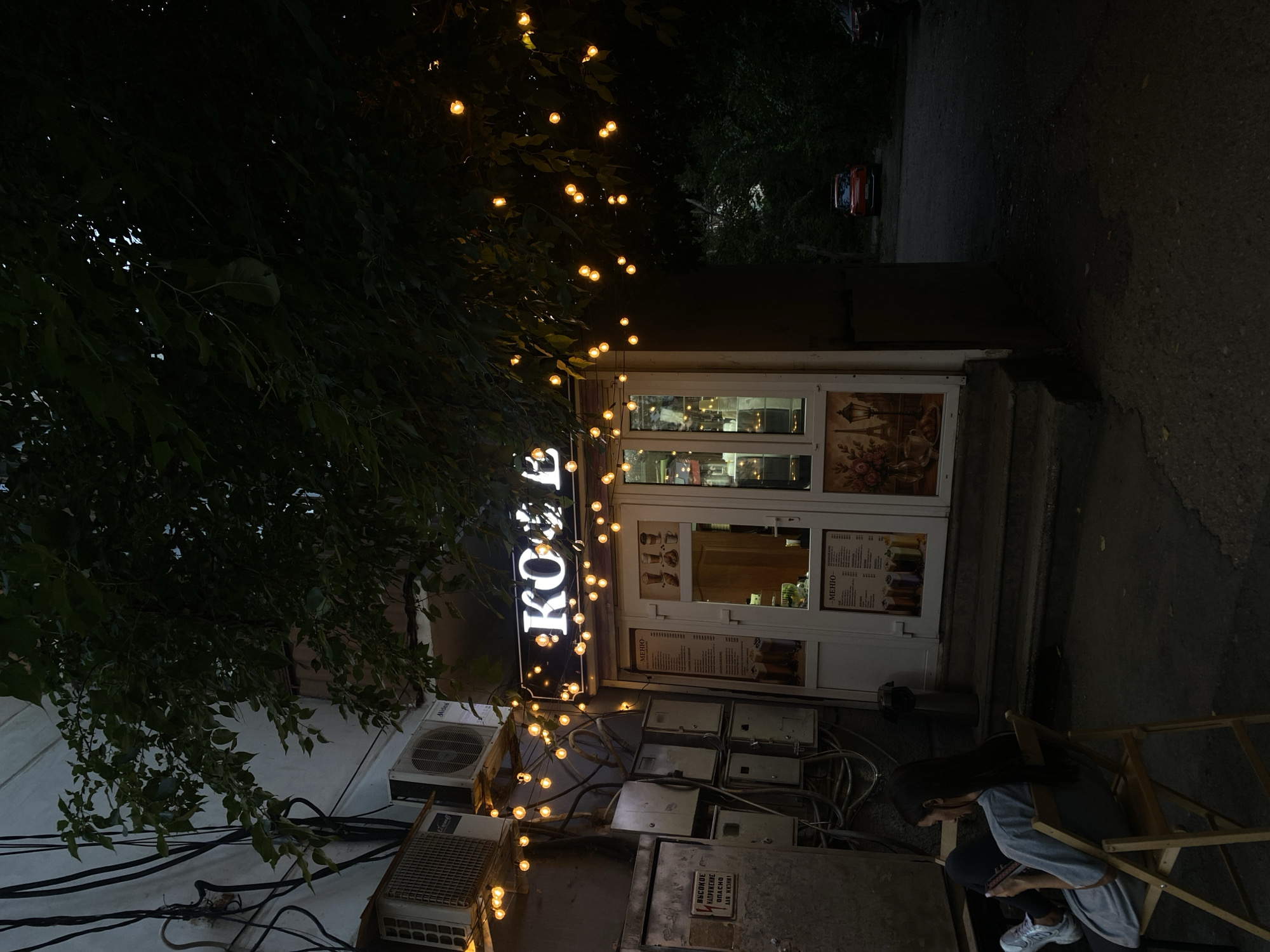 The image depicts a street-level view of a small cafe with illuminated signage reading "KOFE" at dusk in Feodosia, Ukraine. A string of warm-toned fairy lights drapes across the facade of the cafe and extends to the left, illuminating the exterior wall which is adorned with electrical conduits and air conditioning units. To the left of the cafe entrance, a person is seated on a wooden chair, looking down at a mobile device. The cafe features large glass doors with menus posted inside, showcasing beverages and food items. The area around the cafe is paved, and beyond it, trees and a glimpse of a red car are visible. The overall scene is dark, with the cafe's lighting and the fairy lights providing the primary illumination.