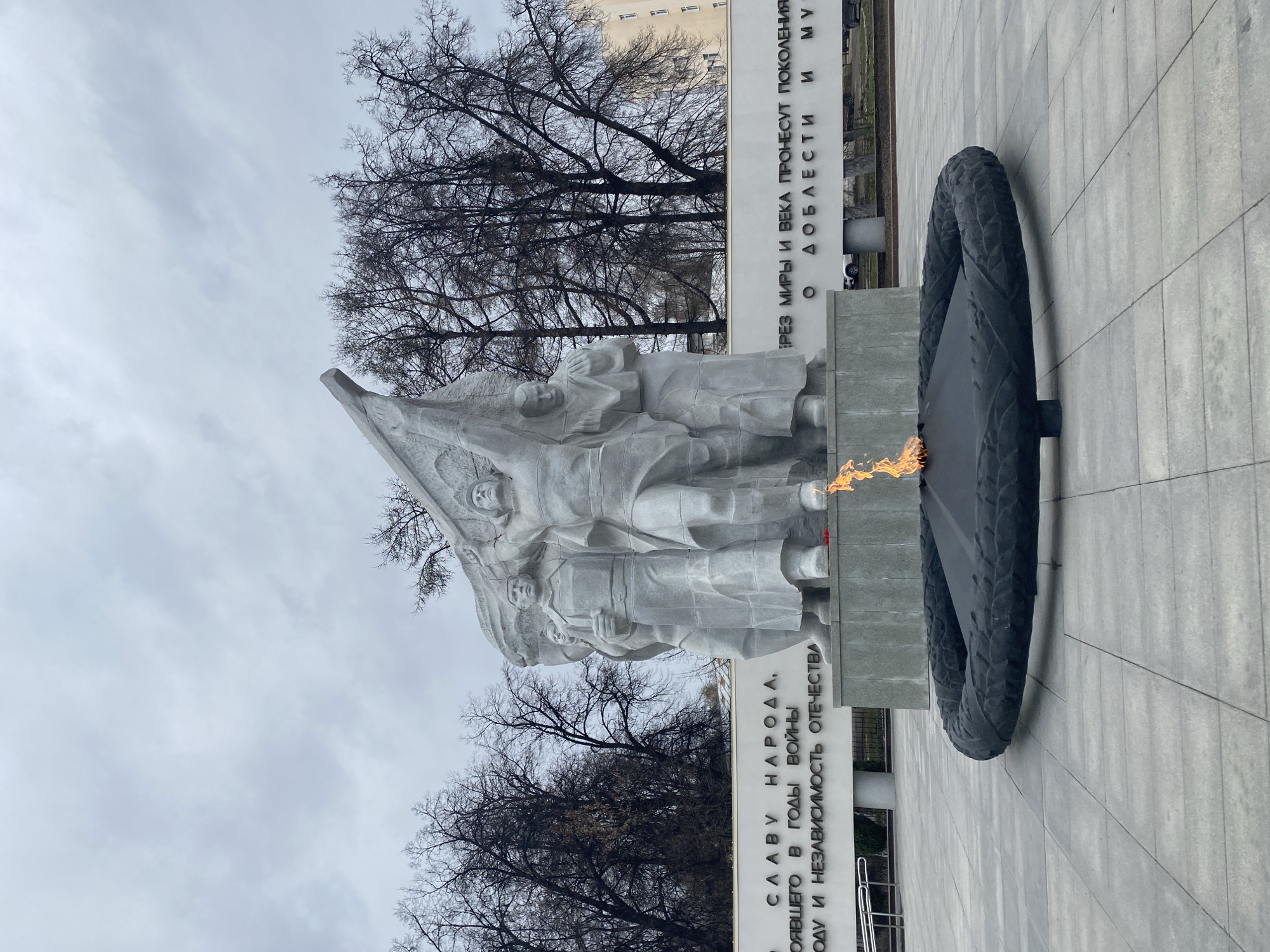 This photograph shows a war memorial in Ryazan, Russia. The monument features a white stone sculpture of several figures holding a banner, next to an eternal flame encased in a dark metal ring. Inscriptions are on the vertical walls behind the statue. Bare trees frame the top of the monument against a cloudy sky. The ground is paved with light gray tiles.