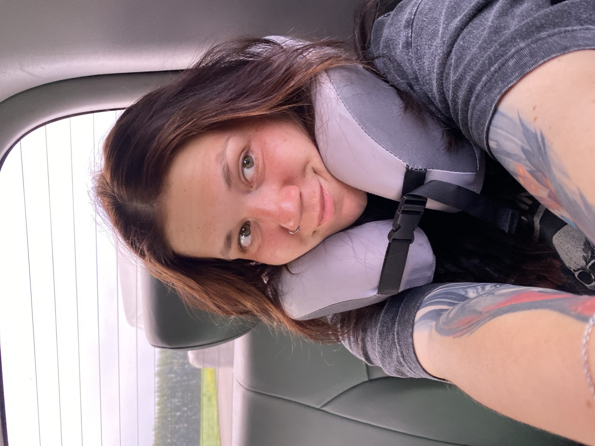A woman with dark brown hair and fair skin is seen from the chest up, leaning against a grey neck pillow. She is wearing a dark grey t-shirt. Her left arm, with colorful tattoos, is visible in the foreground. She has a nose ring and is looking to her right with a slight smile. She is in the back seat of a vehicle, with the headrest of the seat in front of her visible. The background shows a window with a blurry view of trees and a cloudy sky. The image appears to have been taken on a road trip.