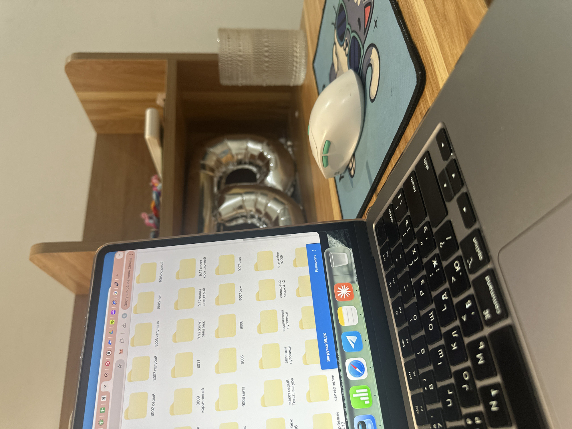The image shows a workstation with a laptop, mouse, and a tablet running a file explorer-like application. The tablet's screen displays folders with names like "8002 серый," "8003 голубой," "8005 лен," and "9003 мята" written in Russian. A chrome update notification is visible. A download is in progress at 99.5%. The laptop keyboard is partially visible, showing Russian characters. A mousepad with a cartoon design is next to the laptop. A shelf above the desk holds a glass and a silver balloon in the shape of the number "8". The photo was probably taken indoors. The precise location is unknown, but the screen showing Russian file names makes it unlikely it was taken in the city of Bac Nha Trang Ward, Vietnam.