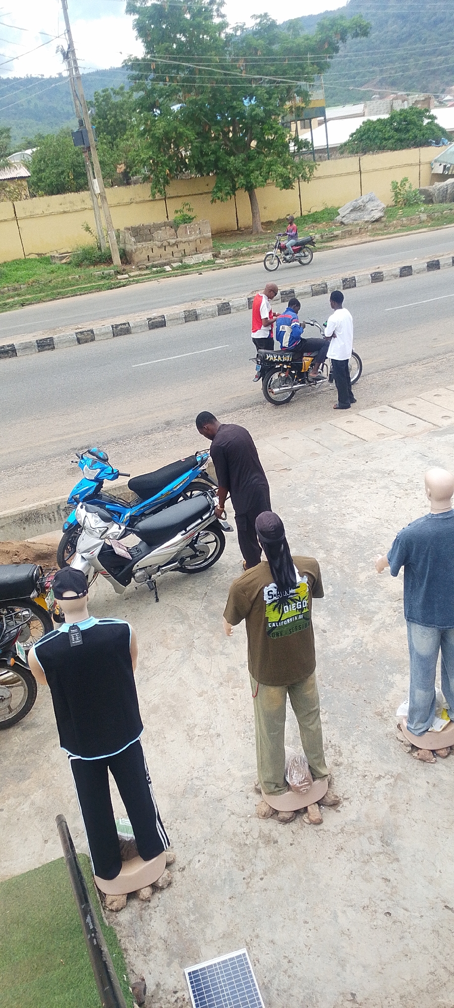 The image depicts an outdoor scene in Lokoja, Nigeria. There are three motorcycles parked, two on the side walk and one on the street, in front of a row of four mannequins. A man in a dark robe bends over one of the motorcycles on the sidewalk. On the street, two men are standing and talking to the driver of the motorcycle. In the background, another person is riding a motorcycle down the street. A small solar panel is on the ground near the mannequins. A yellow wall separates the street from a line of trees and buildings, and a mountain is visible in the background.