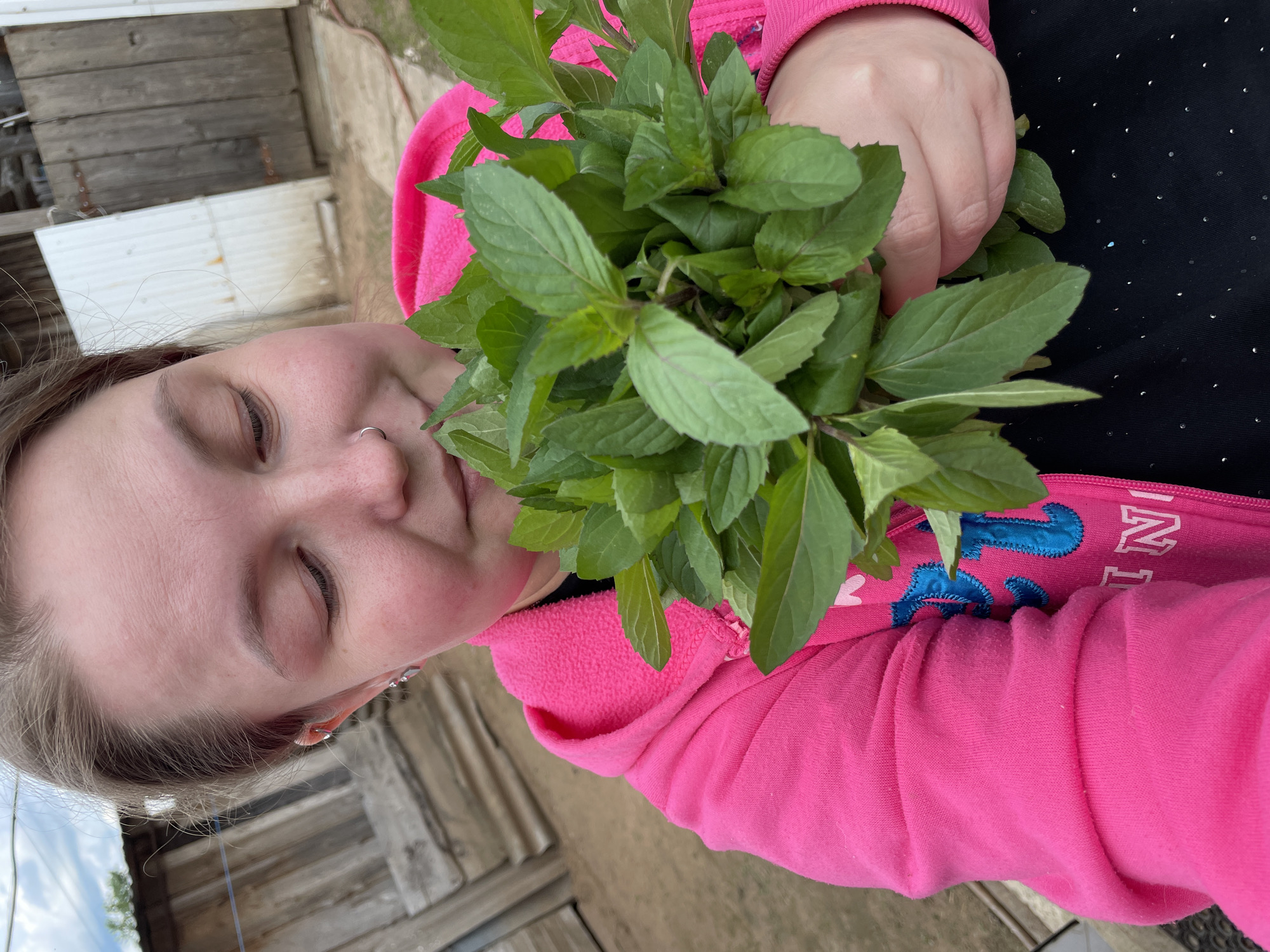 A woman with closed eyes and fair skin is holding a large bunch of leafy green herbs, possibly mint, up to her nose. She is wearing a bright pink zip-up hoodie over a black top with small white dots. The woman has a nose ring and earrings. The background includes a wooden building and possibly a dirt or gravel ground. The image was taken in Yamushan-Kliuchi, Russia.