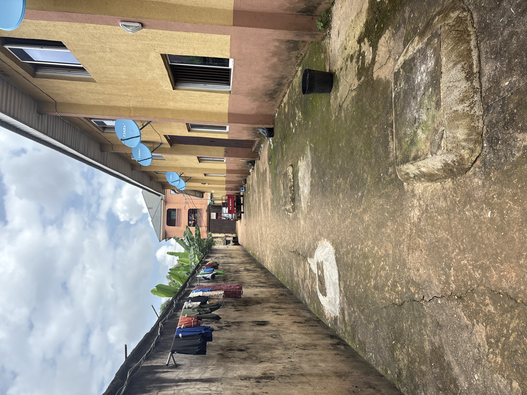 This is a photograph taken in Onitsha, Nigeria. The scene depicts a narrow, outdoor walkway or alley between two buildings. On the left, a textured, aged concrete wall runs the length of the frame, with laundry hanging from a clothesline attached to the wall. The laundry consists of various garments, including shirts, pants, and a skirt. Adjacent to the wall, banana trees are visible, suggesting a tropical or subtropical climate.
On the right side of the alley is a two-story building with a mustard-yellow facade and a brown trim around the base and windows. Several satellite dishes, labeled "DSTV," are mounted on the upper part of this building. The building has multiple windows, some with metal bars for security, and a door visible at ground level.
The walkway itself is paved with concrete, showing signs of wear, including cracks and patches of moss or algae growth. In the foreground, a raised, square concrete structure is visible, also covered in moss. Further down the alley, a small red truck with "FC-MINAJ" written on its side is parked. The sky above is partly cloudy. The overall impression is one of a functional, lived-in urban environment, likely a residential or mixed-use area.