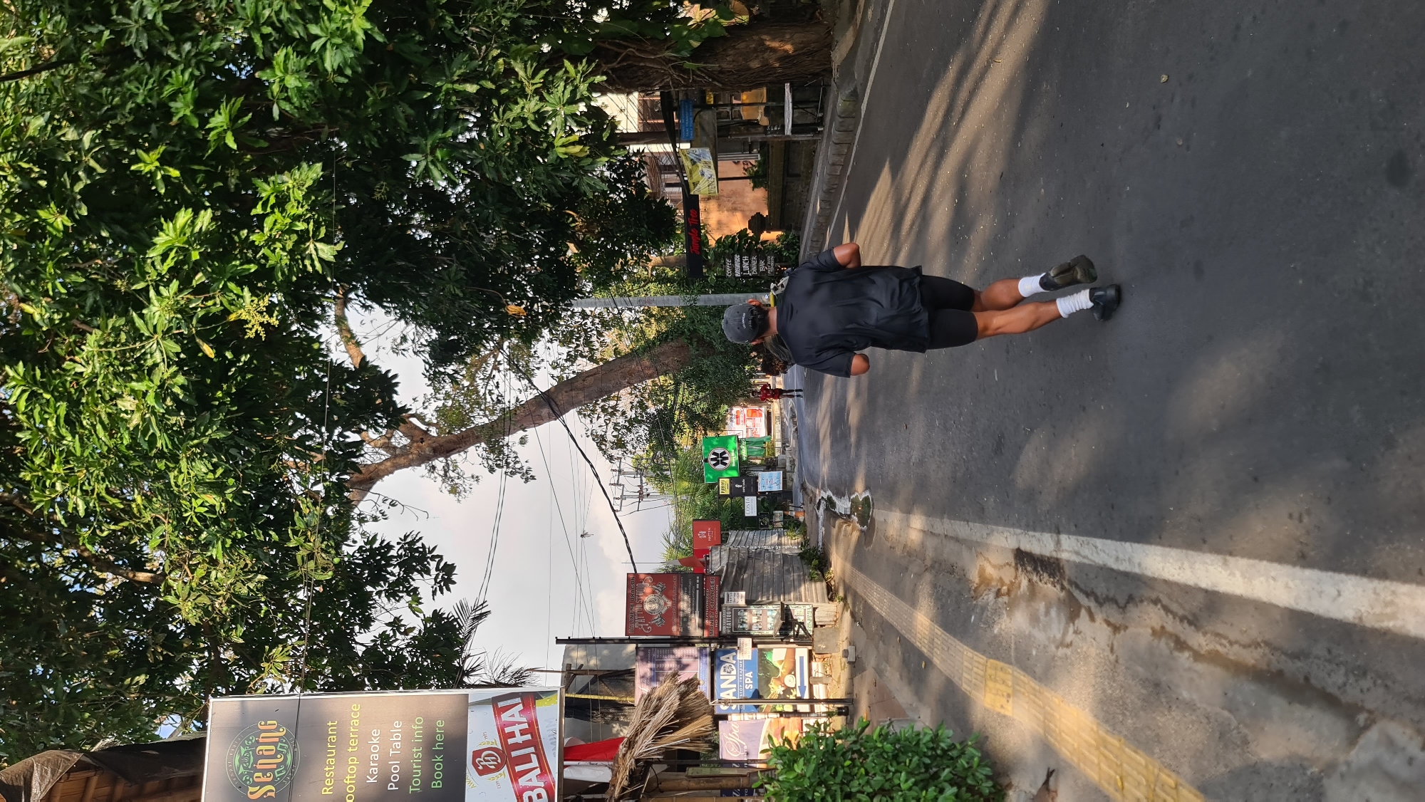 The image depicts a street scene in Denpasar, Indonesia, likely in a tourist area given the signage. In the foreground, a person is jogging away from the camera, wearing a black t-shirt, black shorts, white socks, and black shoes. They are on the right side of a paved road, with a yellow tactile paving strip and a sidewalk on the left. The road has a white line marking the lane.
The environment is characterized by lush green trees overhanging the street. Several signs are visible on the left side of the road, including one that reads "Senang" with smaller text indicating "Restaurant," "Rooftop terrace," "Karaoke," "Pool Table," "Tourist info," and "Book here." Another sign below it says "BALI HAI." Further down the street, more signs for various businesses, including a "Tattoo Studio" and a spa, are visible. In the distance, other pedestrians can be seen, and buildings line the street. The sky is visible through the trees, suggesting it is daytime with overcast conditions.