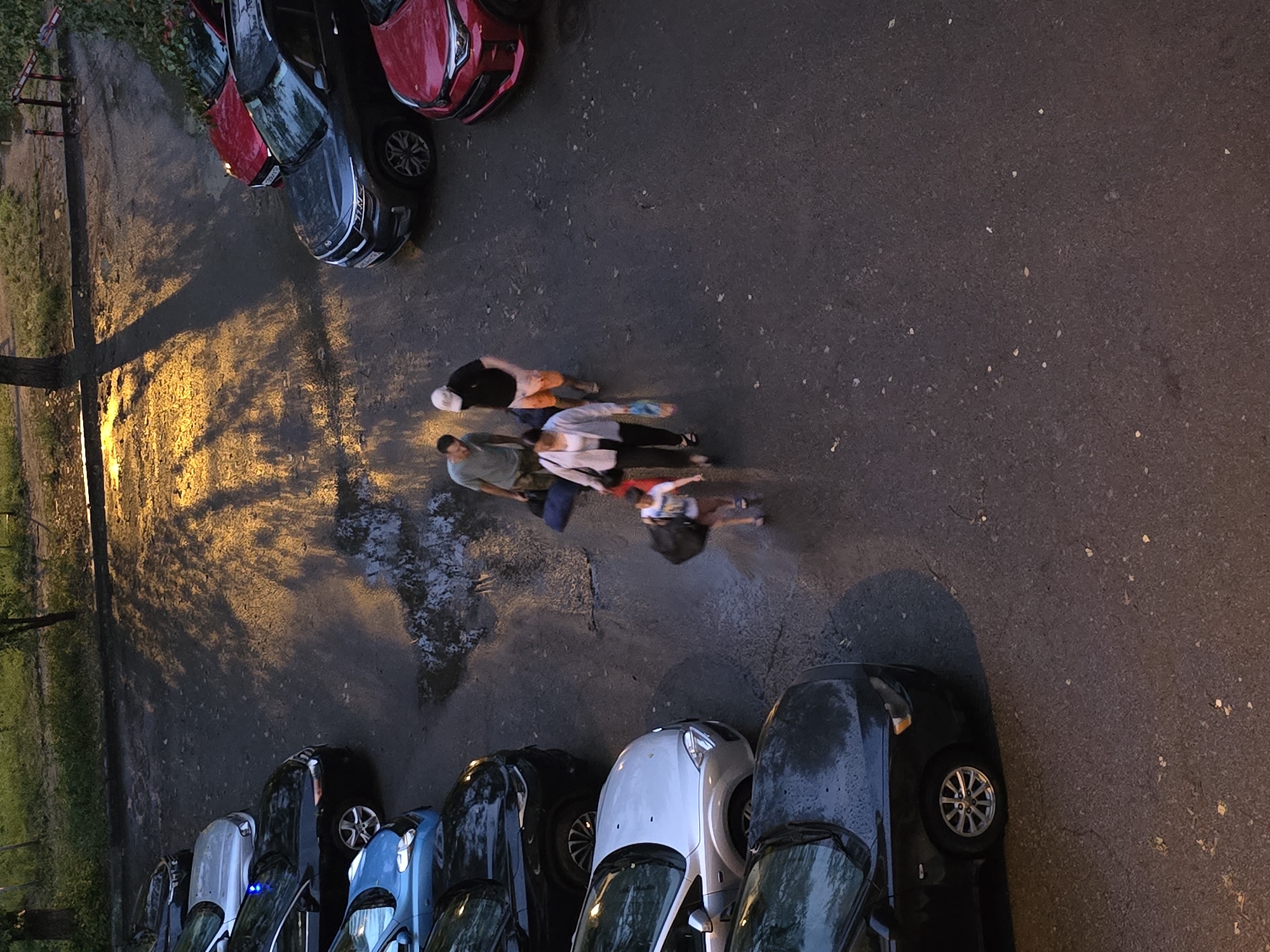This aerial view captures a street scene in Krasnoyarsk, Russia, likely during the evening or at dusk, as indicated by the warm, directional lighting casting long shadows. The ground is wet, suggesting recent rain.
Several parked cars, predominantly dark colors like black and blue, are lined up along the asphalt. One red car is also visible. The cars appear to be in a residential area, possibly a parking lot.
In the middle of the frame, a group of five people are walking together. They appear to be moving from right to left. One person is wearing a white cap. The people are dressed casually in t-shirts and shorts. Their interaction is unclear, but they are in close proximity, suggesting they are together. The overall atmosphere seems ordinary, with no overt signs of distress or urgency. The location cues, like the cars and the general urban setting, are consistent with a Russian city.