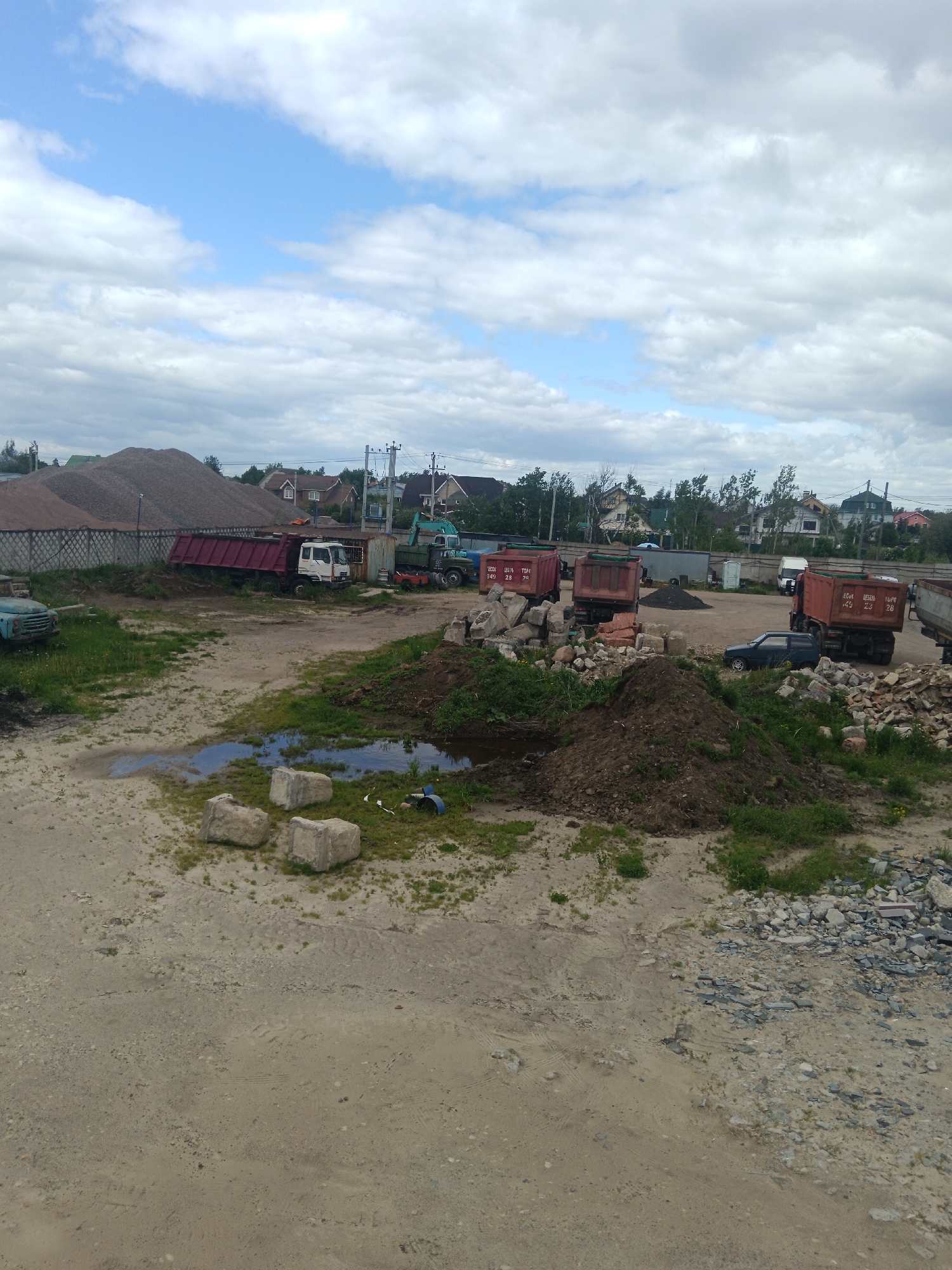 The image shows a construction or industrial site in Яльгелево, Russia. In the background, there are residential buildings and a large pile of gravel or sand. Several trucks, some with red dump beds, are parked in the area. A blue car is also visible. In the foreground, there's a dirt area with puddles, piles of dirt, grass, and scattered concrete blocks and rocks. The sky is cloudy.