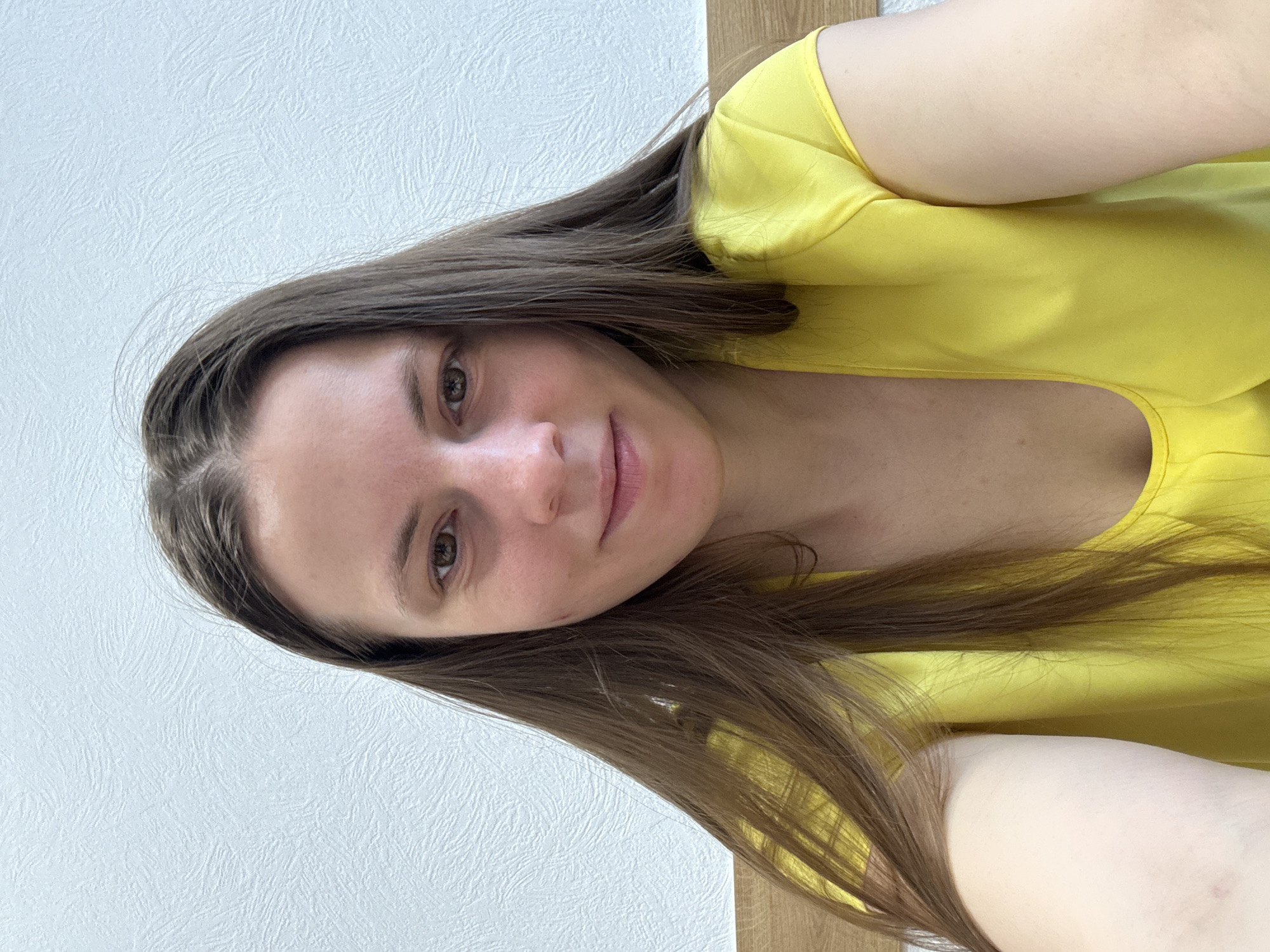 A medium close-up selfie captures a young woman with long, straight, dark brown hair that frames her face and extends over her shoulders. She has a light complexion, brown eyes, and a subtle, pleasant smile. She is wearing a bright yellow top or dress.
The setting appears to be indoors, with a textured white wall visible behind her head and upper body. To the right side of the frame, a light brown wooden surface, possibly furniture or a headboard, is visible behind her lower body and arm. The lighting is soft and even, suggesting daytime conditions indoors. There is no visible text in the image.
The woman is looking directly at the camera, indicating she is taking the photograph herself. Her expression is calm and gentle. While the image itself does not provide explicit location cues, the user specified the context as Chaikovsky, Russia.