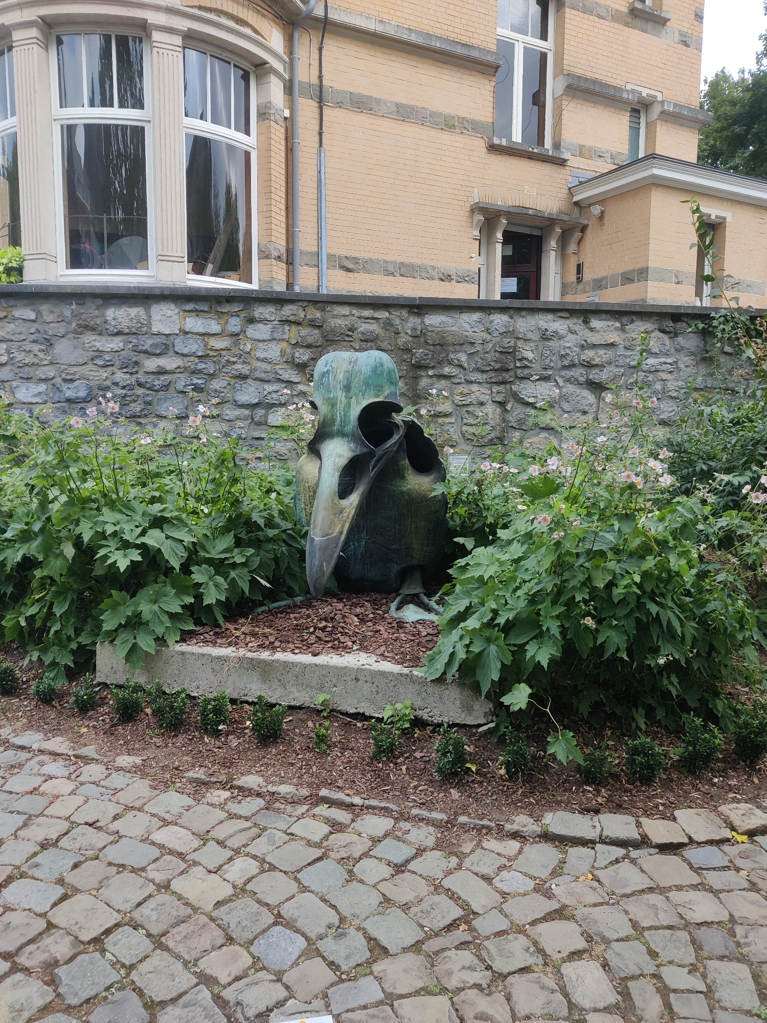 The image shows an outdoor scene in front of a building with a light yellow brick facade and large windows. In the foreground, a cobblestone path leads to a garden bed with a stone retaining wall. A large, abstract metal sculpture resembling a bird's skull sits in the garden, surrounded by lush green plants and some small pink flowers. The building appears to be historic, with architectural details such as columns and stone accents. The sky is not visible in the frame. The setting suggests a public space, possibly a park or a courtyard adjacent to a building.