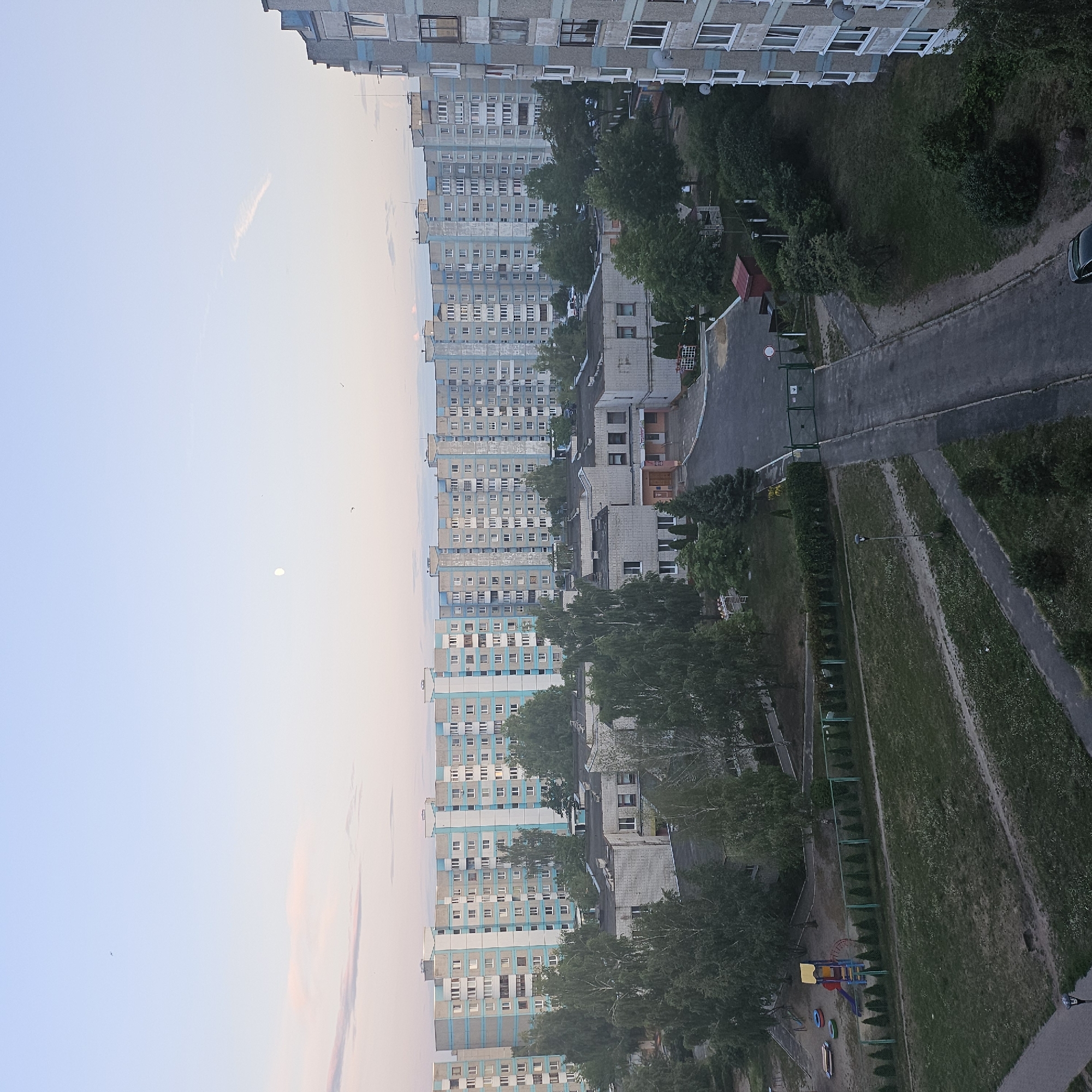 The image depicts an aerial view of a residential area in Brest, Belarus, during twilight. In the foreground, a playground with a slide, swings, and sandpit is visible. Adjacent to the playground is a patch of grass and a fence. Several trees and shrubs are scattered throughout the scene. In the middle ground, a low-rise building with a gray facade and a dark roof is present. Further in the background, a row of tall, multi-story apartment buildings with light blue and gray facades stretches across the horizon. The sky is a soft gradient of light blue and pink, with a faint crescent moon visible and a few wispy clouds. The overall impression is a quiet, early morning or late evening in a typical urban environment. There are no people or animals visible in the image.
