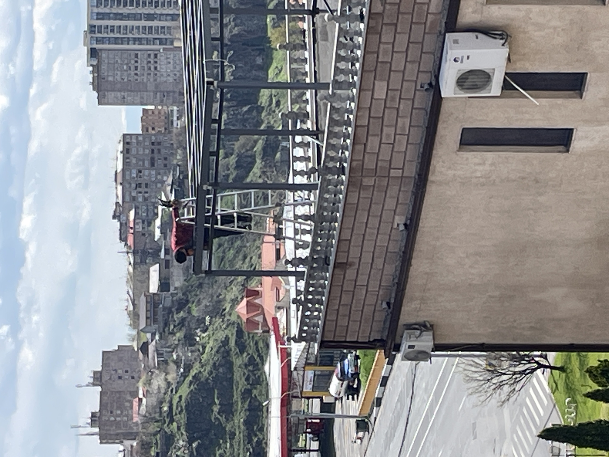 An elevated view in Yerevan, Armenia, looking southwest across the city.
In the foreground is a building with a tiled roof and an air conditioning unit mounted on the wall. A metal scaffolding structure is positioned in front of the building's edge, with a worker visible on the scaffolding, wearing a red shirt and black pants, likely performing maintenance or construction work.
Beyond the building, a street runs parallel, featuring a white vehicle, traffic lights, and trees. The background reveals the urban landscape of Yerevan, with residential buildings and dense greenery covering the hillsides. The sky is cloudy.
