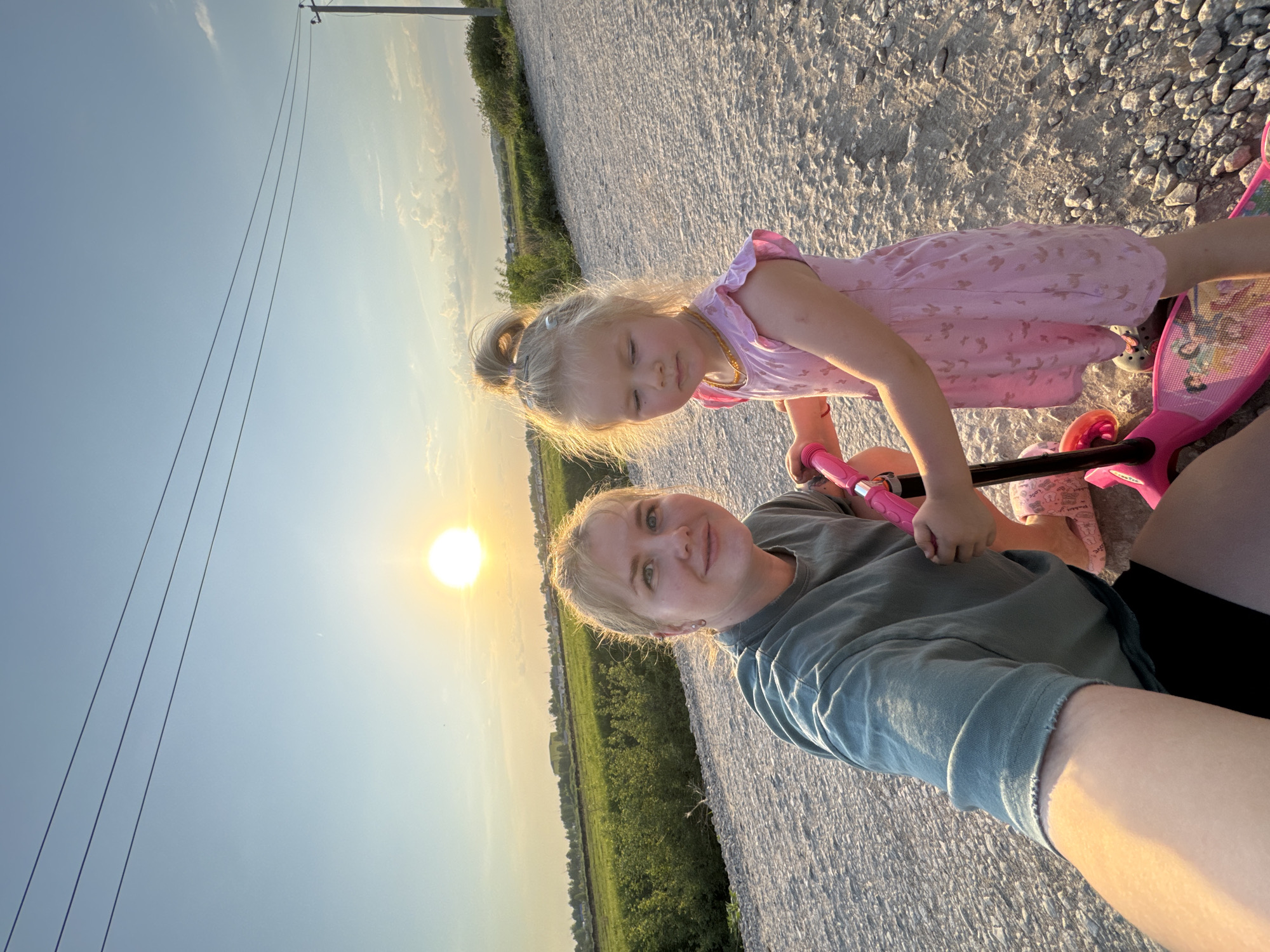 A selfie is taken outdoors, with a fair-skinned adult and a fair-skinned child on a pink scooter. The sun is positioned behind them in a clear sky. The location appears to be a gravel road near Горный, Russia. The adult is wearing a green shirt and black shorts. The child has blond hair in a ponytail and is wearing a pink dress. The ground is composed of light-colored gravel. Power lines are visible in the background.