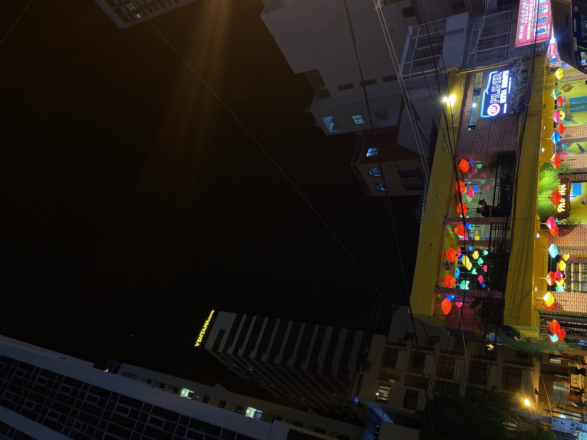 This is a night shot of a street scene in Nha Trang Ward, Vietnam, with buildings and colorful decorations. The image is rotated, with the buildings appearing to hang from the top. A prominent feature is a building with a yellow facade adorned with numerous colorful lanterns, hanging from the top of the building and along the street. Some of these lanterns are illuminated, casting a vibrant glow.
On the yellow building, a neon sign reads "Pilsner" in blue and red, with "Erotro" underneath. To the right, another sign indicates "MINI MART - ĐỒ GỖ MỸ NGHỆ T". Further down the street, more buildings are visible, some with illuminated windows. One building on the left has a large, illuminated sign that reads "VENTANA HOTEL".
People can be seen at tables on the ground floor of the yellow building, suggesting a restaurant or bar is in operation. The overall atmosphere appears lively and festive, with the bright lanterns creating a cheerful ambiance against the dark night sky. The scene is urban, likely a commercial street or tourist area given the presence of hotels, shops, and illuminated signs. The time of day is clearly night, with artificial lighting playing a significant role in illuminating the scene.