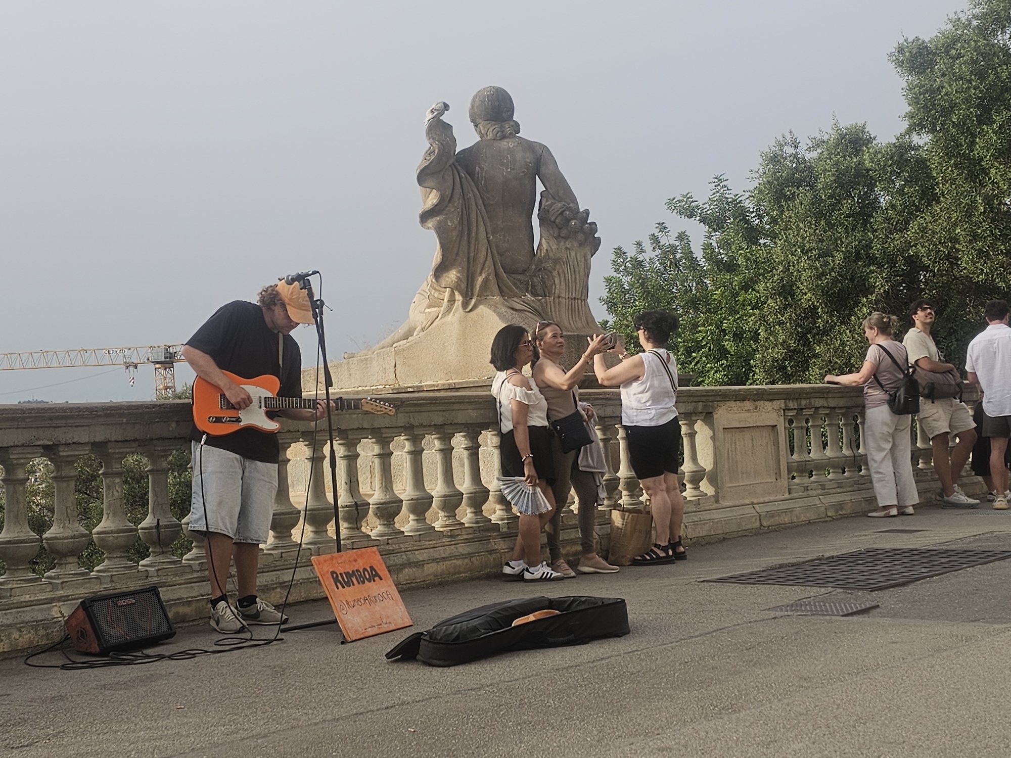 The image captures an outdoor scene, likely in Barcelona, Spain, given the context of public art and street performers often found there. In the foreground, a man in a black t-shirt and light-colored shorts is playing an orange electric guitar. He wears an orange baseball cap and is positioned in front of a small amplifier. To his right, an orange sign with the word "RUMBOA" and a social media handle is placed on the ground. A black guitar case lies on the pavement near the sign. Behind the musician, a stone balustrade separates the paved area from a higher level where a large stone statue stands. The statue depicts a figure with draped clothing and appears to be a classical sculpture. A group of people are visible near the statue. Three women are taking photos with their phones. Two of them are wearing light-colored tops and dark skirts, while the third woman is in a white top and dark skirt. Further to the right, a man and a woman are standing, the man in a white shirt and the woman in a white top. In the background, lush green trees and the sky form the backdrop, with a construction crane visible in the distance, suggesting ongoing development or infrastructure work in the city. The overall atmosphere suggests a public space where art, music, and leisure activities converge.