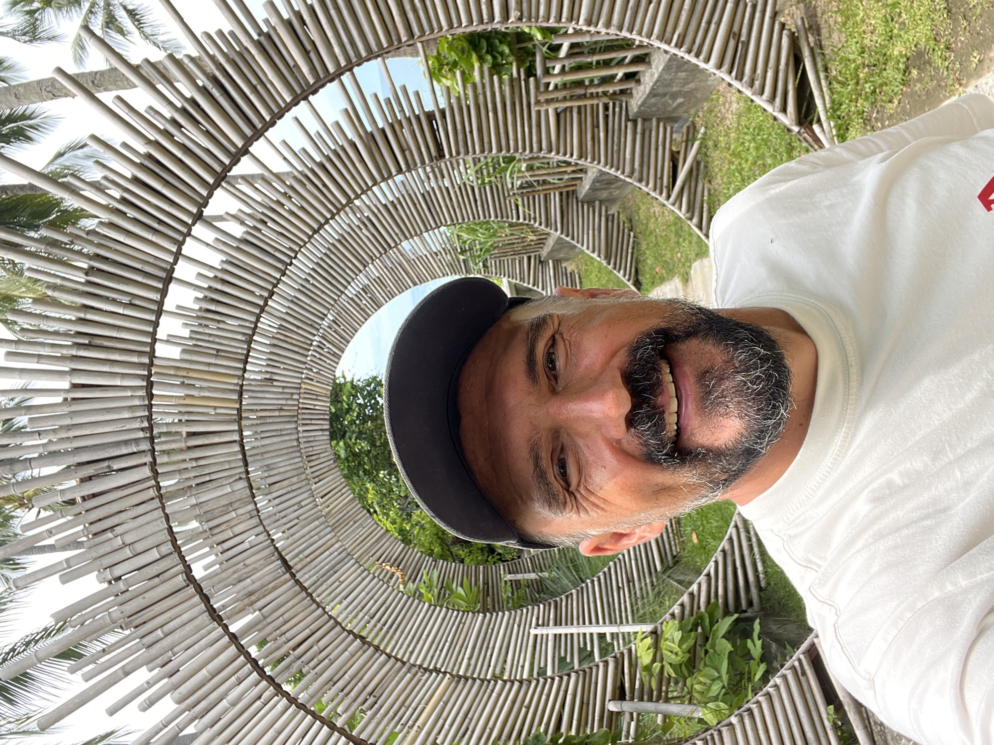 A man wearing a dark baseball cap and a white t-shirt takes a selfie in front of a circular bamboo structure. The man has short, dark hair and a salt-and-pepper beard and mustache. He is smiling and looking at the camera. The bamboo structure is made of many thin bamboo poles arranged in concentric circles, creating a tunnel-like effect. Behind the man and the bamboo structure, there are palm trees and lush green vegetation. The overall setting appears to be outdoors in a tropical environment, possibly a garden or a resort. This scene is located in Baan Chaweng Noi, Thailand.
