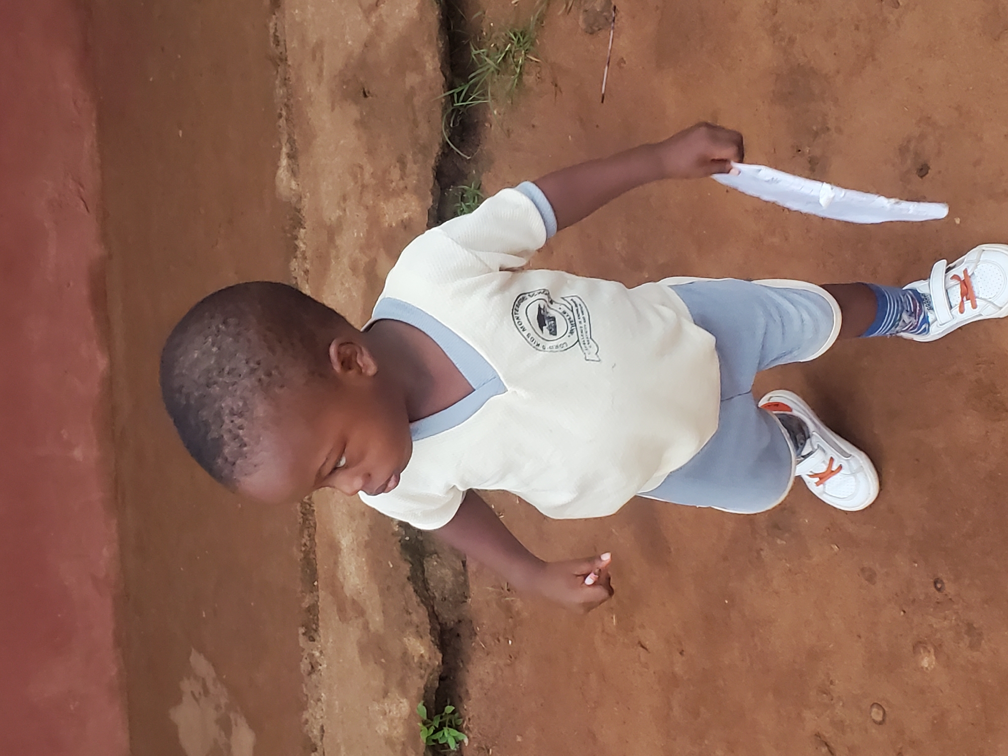 A young child stands on a bare dirt surface adjacent to a wall, holding a white cloth object. The child wears a light-colored shirt with a dark trim, shorts, striped socks, and white shoes with orange accents. The location is possibly Ukwu-Abwa, Nigeria, although this cannot be forensically verified from the image alone. The child appears to be looking down. There are some weeds near the wall. The bare dirt surface contains small pebbles.